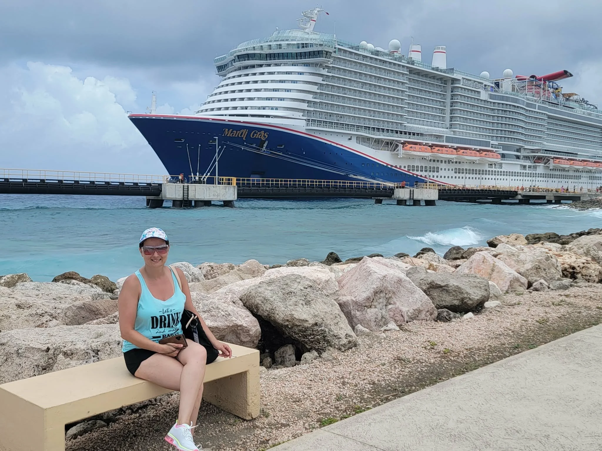 A woman sitting on a bench near the rocky shoreline, smiling and holding a smartphone, with a large cruise ship named "Mardi Gras" docked in the background under cloudy skies.