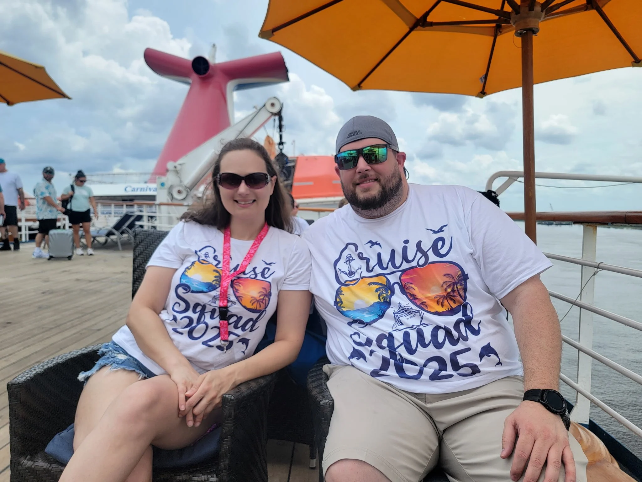 A smiling couple wearing matching white shirts with colorful sunset and palm tree designs, sitting on a boat deck under orange umbrellas, with a cruise ship in the background and people walking nearby.