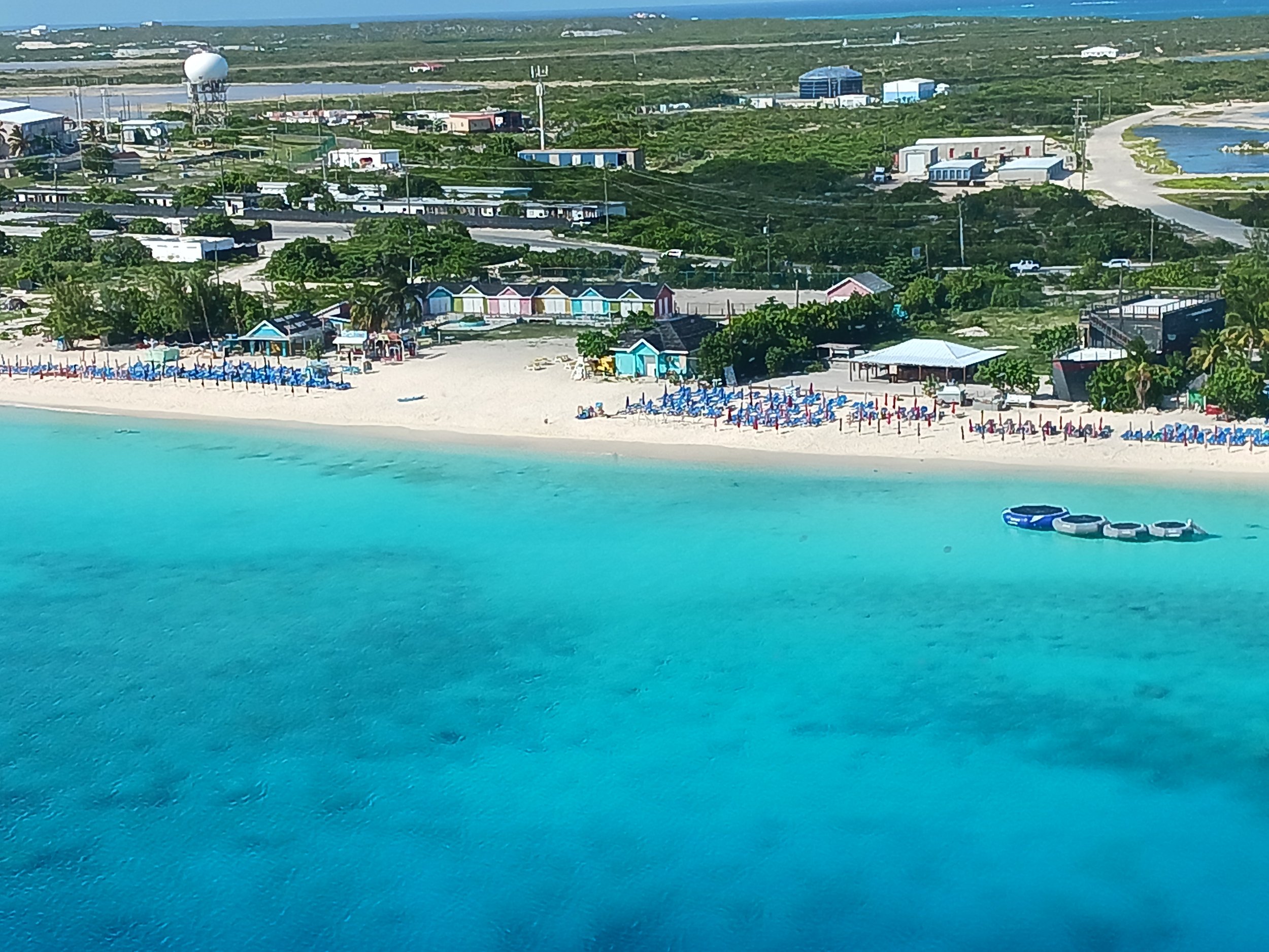 Aerial view of a beachfront area with colorful beach huts, lounge chairs, and umbrellas along a sandy beach, with turquoise water in the foreground and green landscape with buildings and roads in the background.