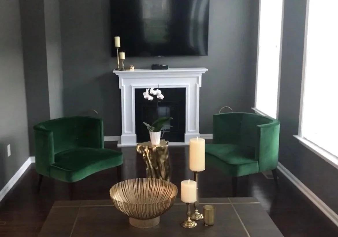 Living room with dark gray walls, a white fireplace mantel, two green velvet chairs, a small gold side table, and candle holders with candles on a dining table. There is a large flat-screen TV above the fireplace and windows with white blinds on the 