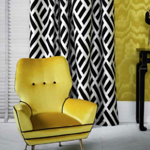 A yellow armchair with black legs in front of a black and white patterned curtain, with a yellow wall and black table in the background.