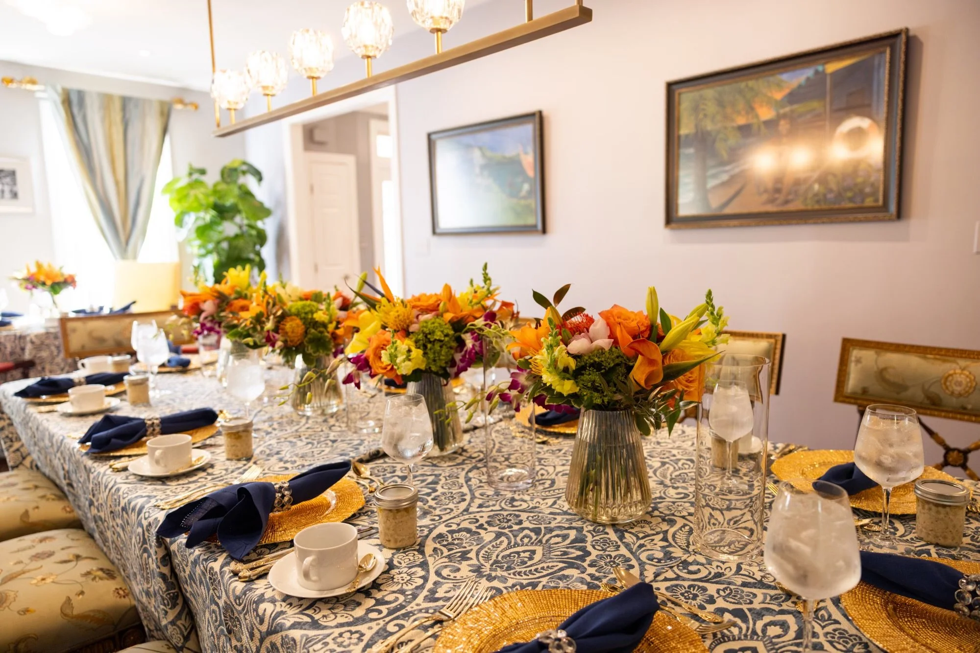 A decorated dining table with a blue and white patterned tablecloth, floral centerpieces, glassware, and place settings, in a well-lit room with framed paintings and a hanging light fixture.