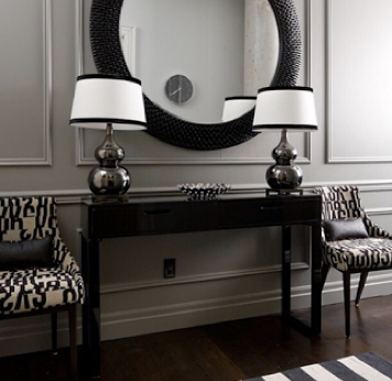 A hallway with a black console table, two patterned chairs, a large circular mirror, and table lamps, in a stylish home interior.