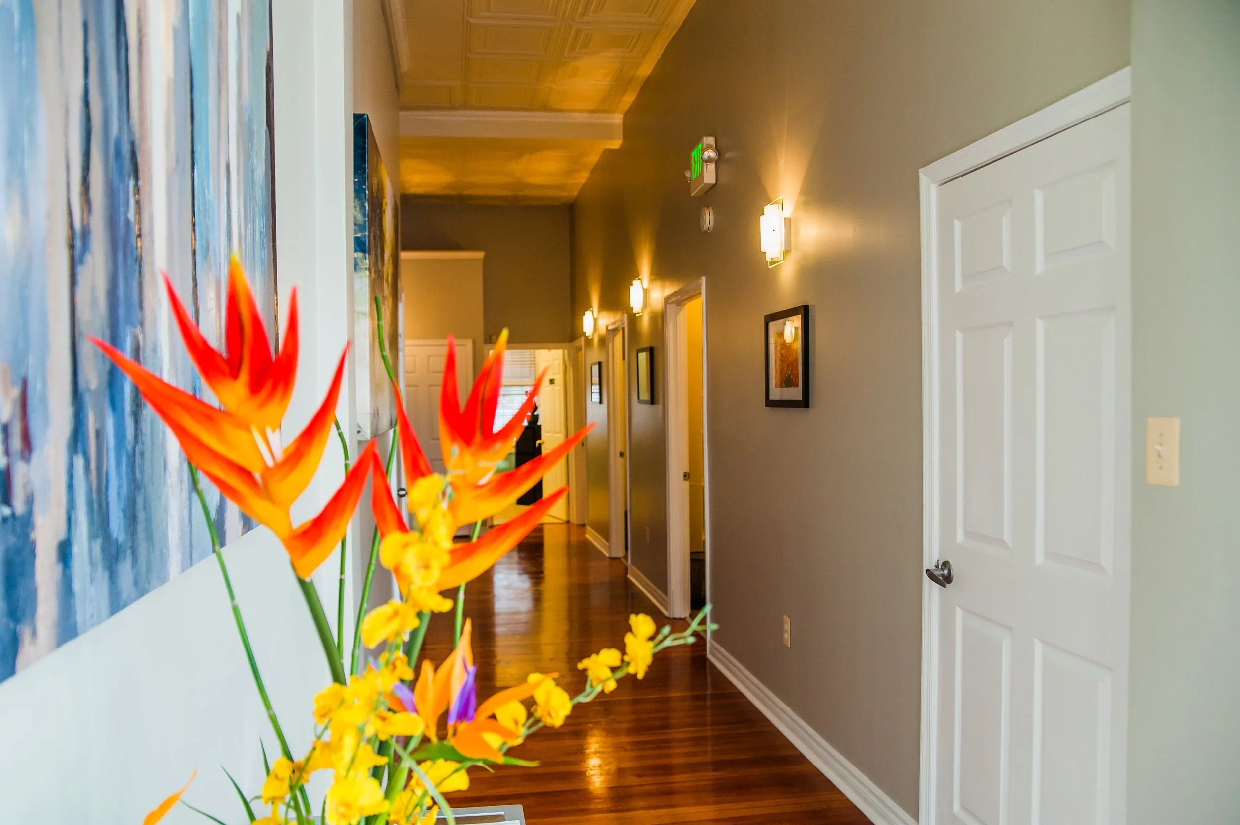 An interior hallway with wooden floors, gray walls, and white doors. There are framed pictures on the wall, bright wall-mounted lights, and a large painting. In the foreground, there is a vase with orange and yellow flowers.