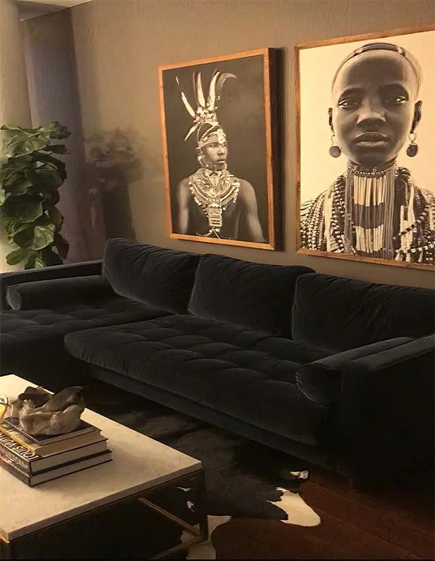 A black velvet sofa in a living room with two black and white portrait frames of African women hanging on the wall behind it, with a plant on the left side and a coffee table with books and decorative objects in front.