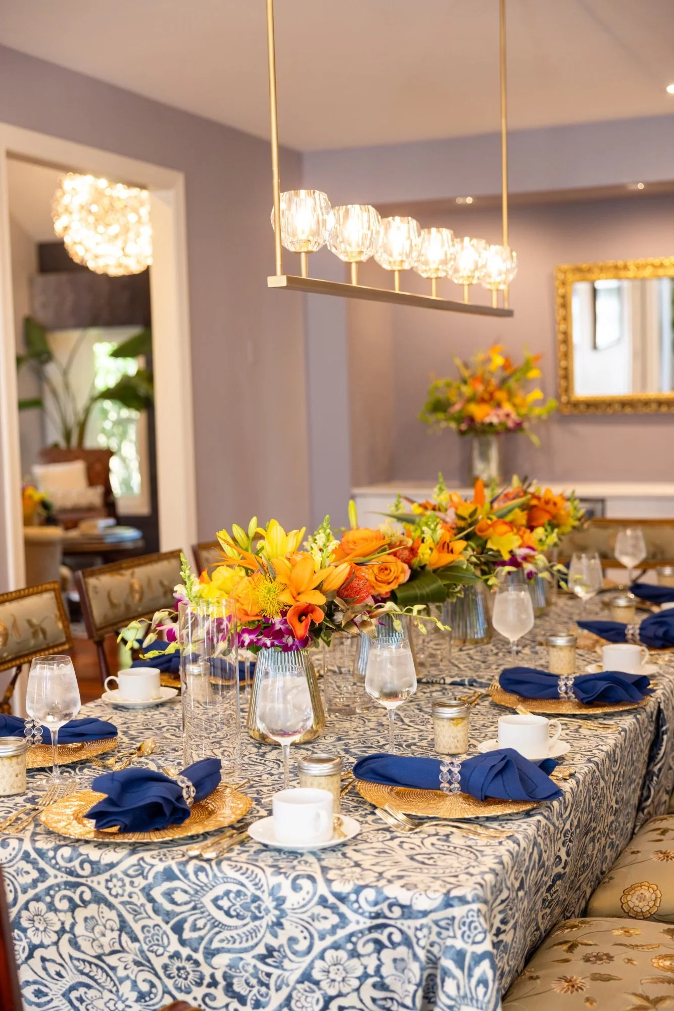 A dining table set for a formal meal with a blue and white patterned tablecloth, floral centerpieces, blue napkins, gold placemats, and glassware in a decorated room with a chandelier and a mirror.