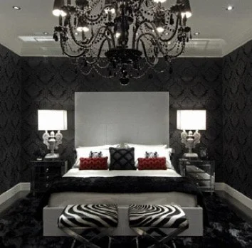 Elegant bedroom with black patterned walls, a black chandelier, and a white bed with red and black pillows, flanked by two nightstands with lamps.