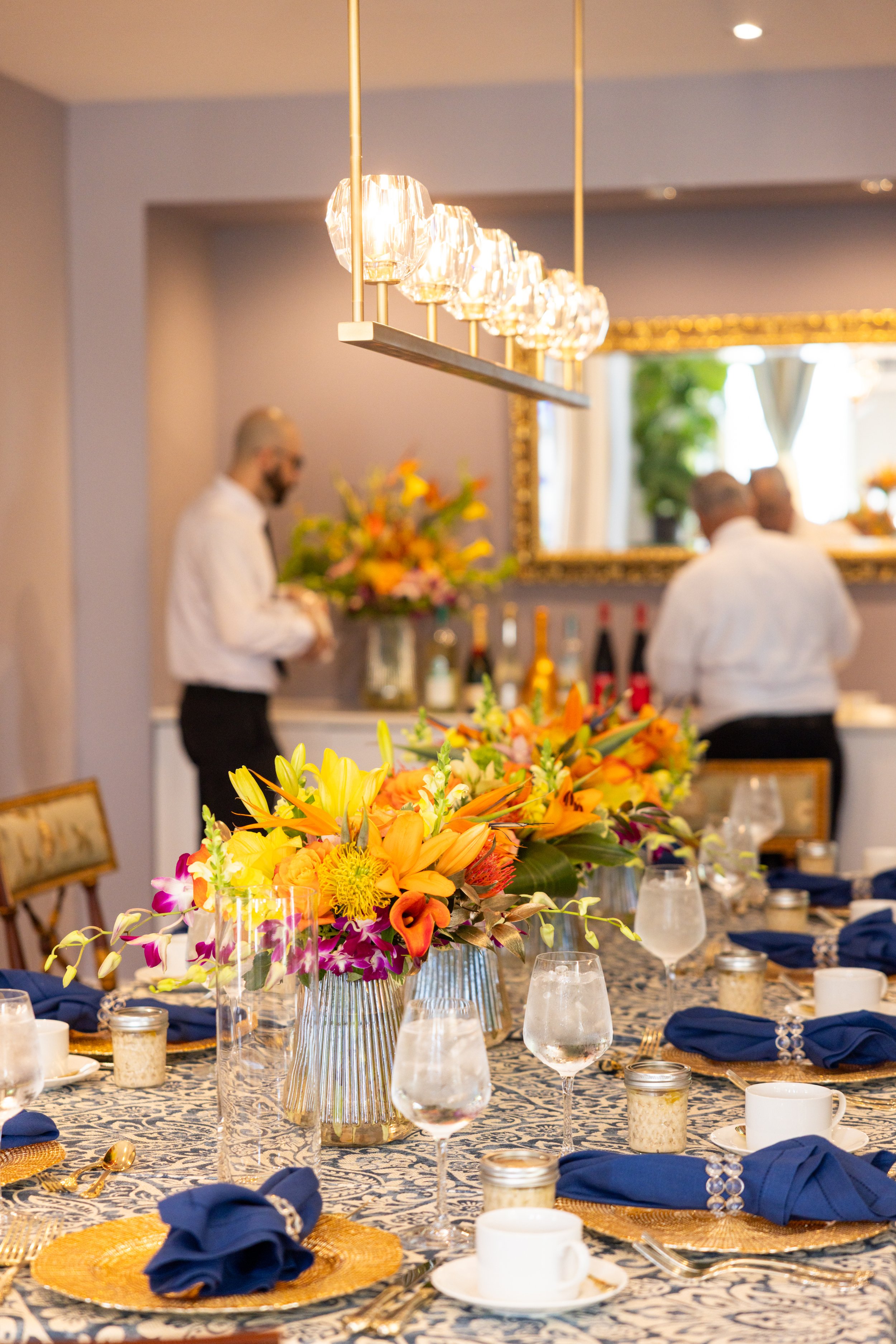 A decorated dining table with a large colorful floral centerpiece, blue napkins wrapped with silver rings, gold placemats, glassware, and candles. In the background, two men in white shirts are talking near a bar with liquor bottles and a large mirror.