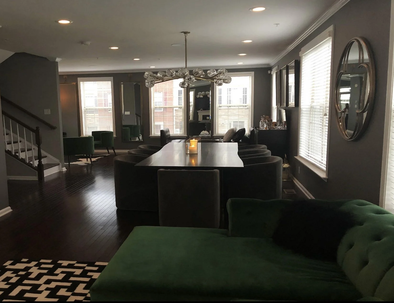 Living room with dark wood flooring, gray walls, large windows with white blinds, and modern decor. A long table is surrounded by velvet chairs, and a green sofa is in the foreground. A chandelier hangs above the table, and candles are lit on the tab