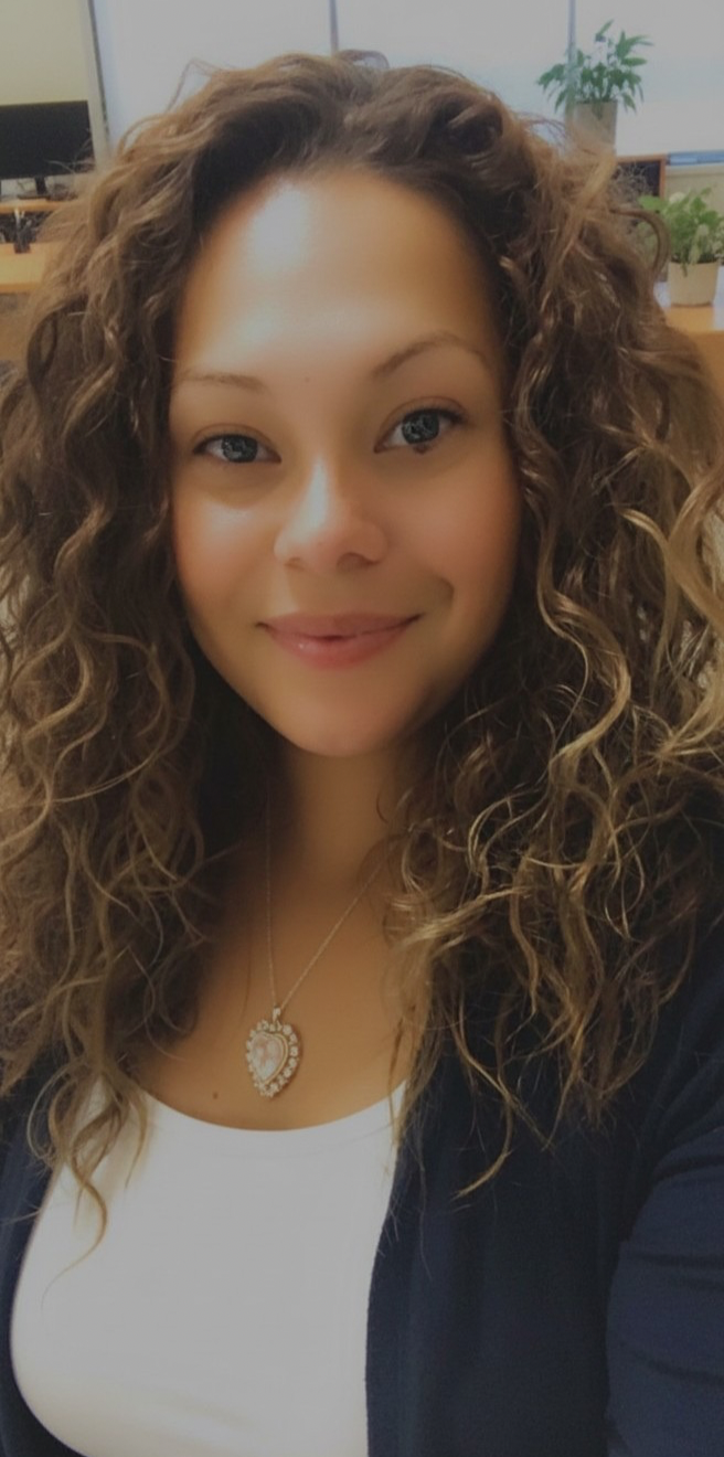 A woman with long, curly, light brown hair smiling in an indoor setting with plants and office furniture in the background.