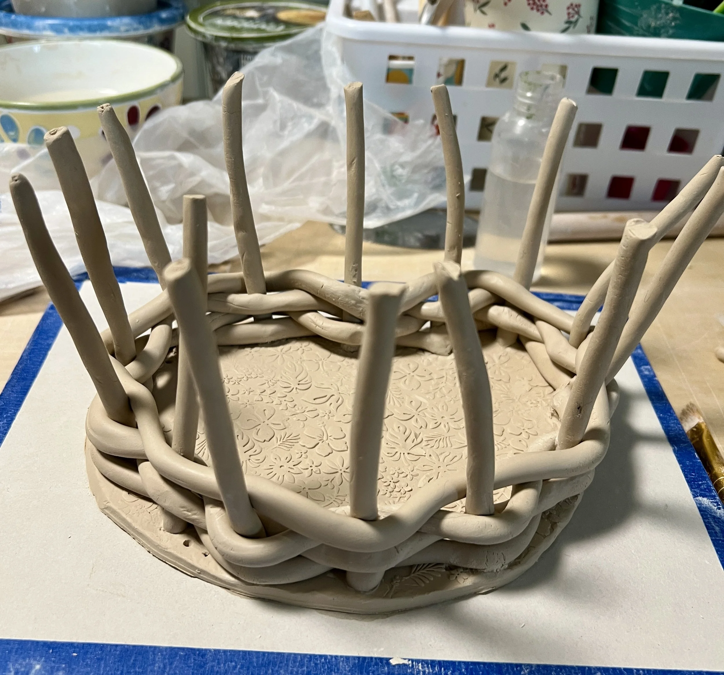 Close-up of a white decorative ceramic basket with a woven design, placed on a table with a textured doily underneath. In the background, there are various kitchenware items including bowls, a plastic container, and a small glass bottle.