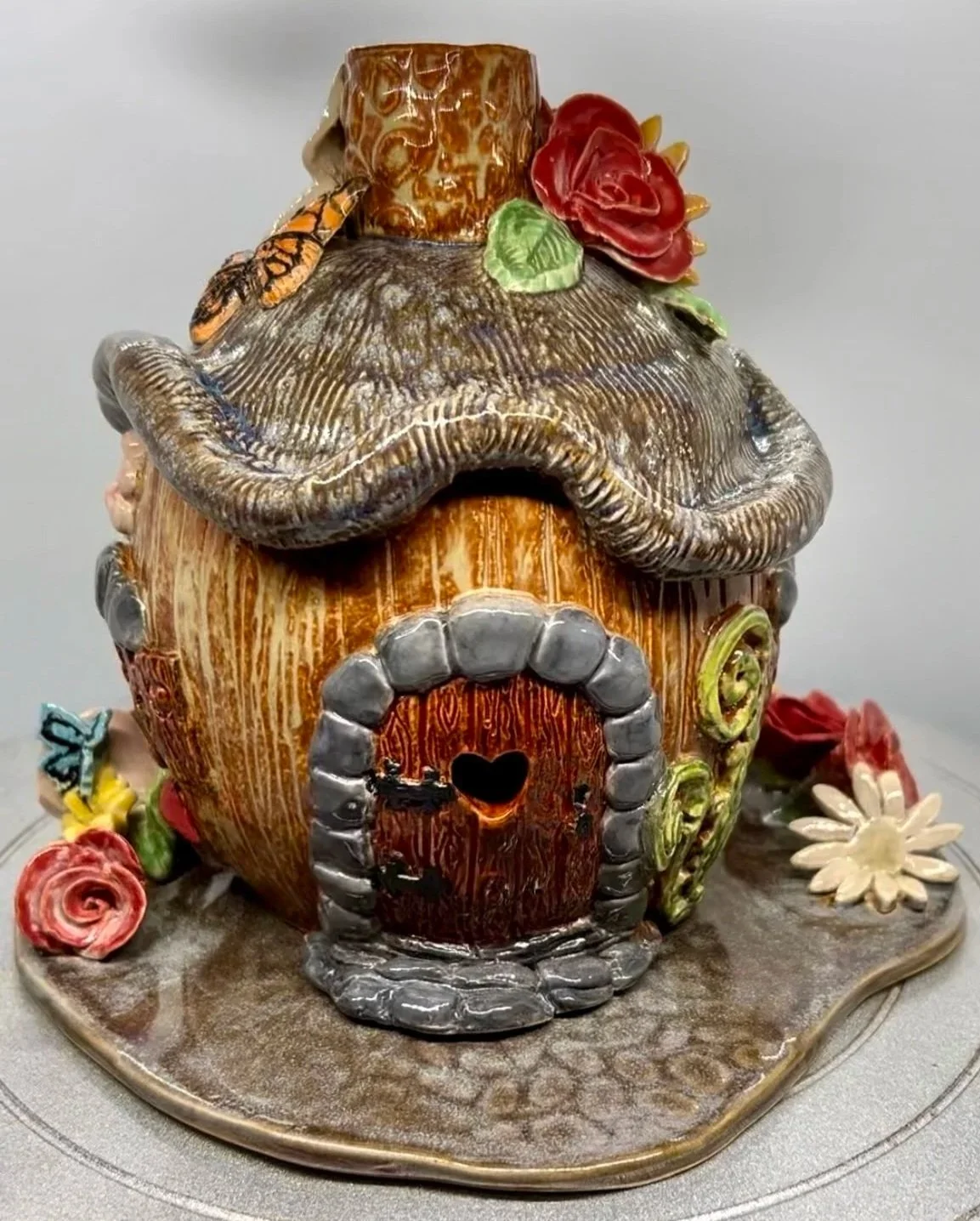 Decorative fairy house with rounded  roof, colorful flowers, a butterfly, and a rose, with a heart-shaped cutout in the door.