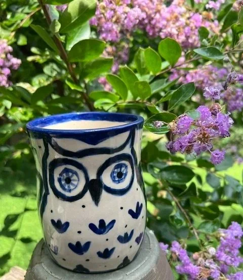 Ceramic owl-shaped cup with blue and white design placed on a stone surface next to purple flowering bushes.