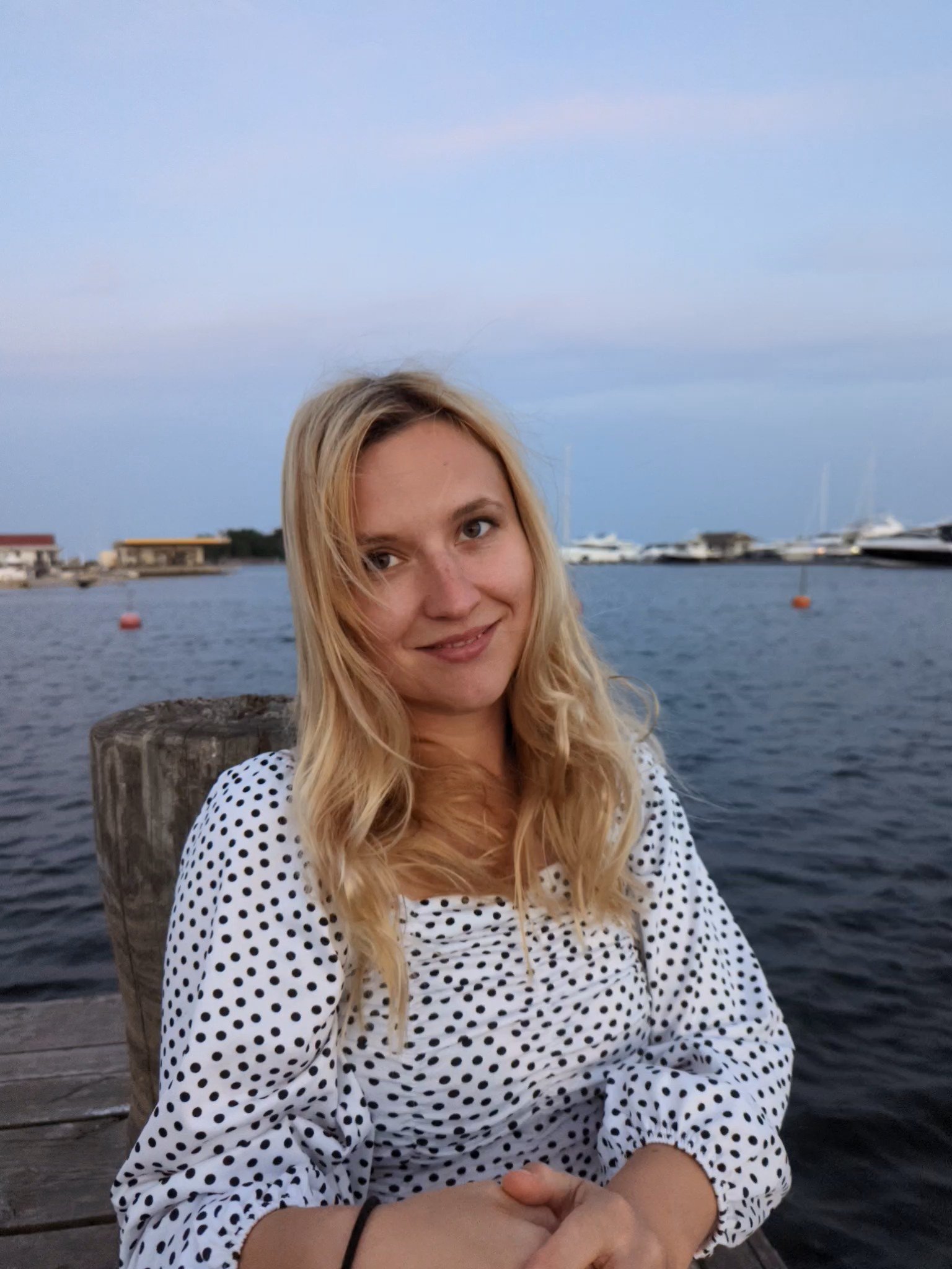 A young woman with long blonde hair, wearing a white dress with black polka dots, sitting by the water at a marina with yachts and boats in the background.