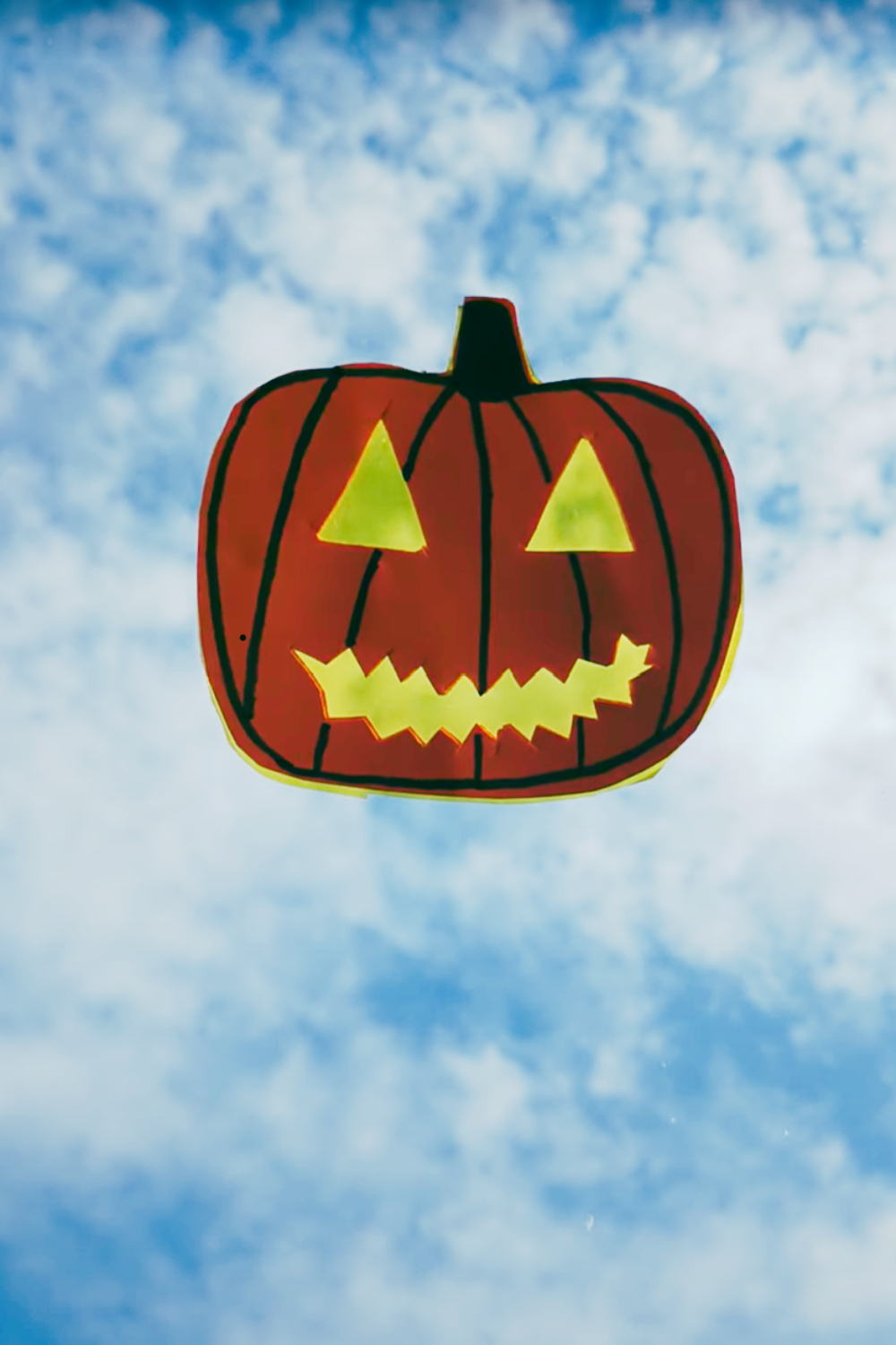 Finished craft: Handmade orange pumpkin suncatcher on a window against a blue, cloudy sky, with light shining through its yellow triangular eyes and zigzag smile.