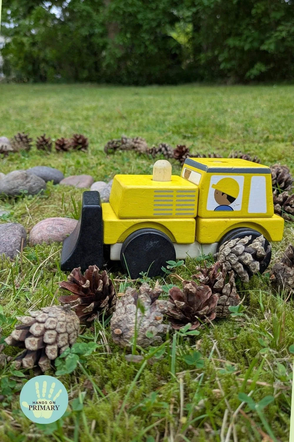 Yellow toy construction vehicle on a natural outdoor racetrack made from stones and pine cones in the grass.