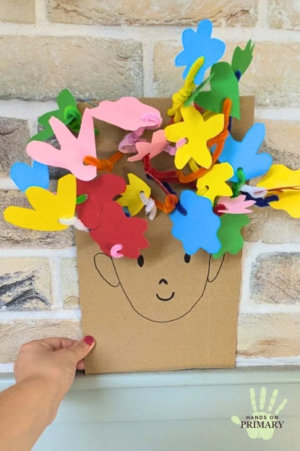 Cardboard and paper craft of a boys face, with flowers in hair made by cutting coloured paper flower shapes and attaching them with pipe cleaners.