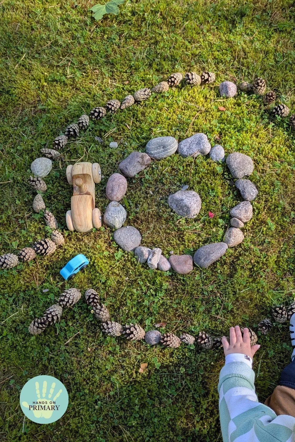 Child playing with toy cars on an outdoor racetrack made from stones and pine cones arranged on the grass.