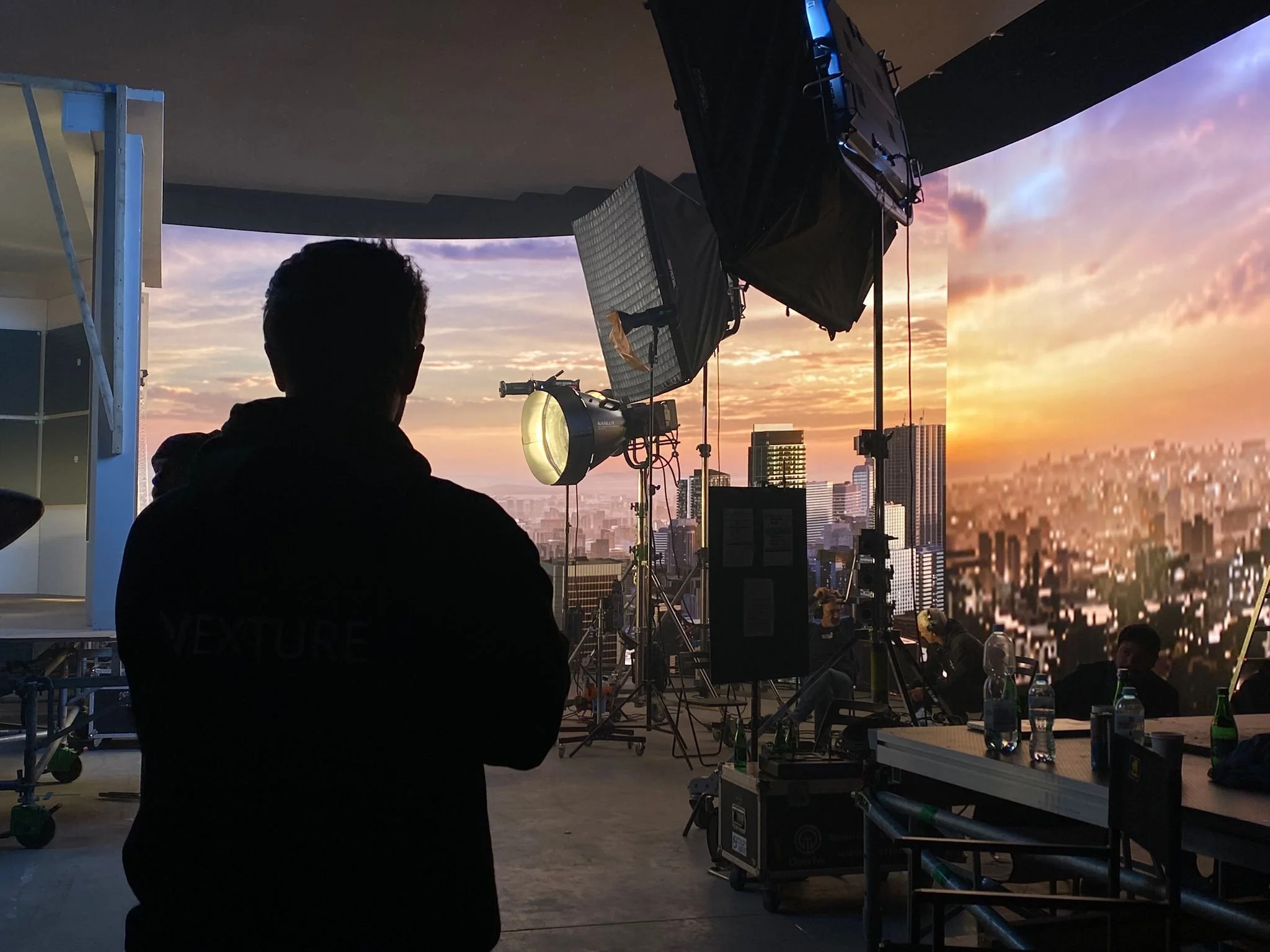 A set scene with film or photography equipment set up inside a building, overlooking a cityscape at sunset through large windows, with some people sitting and working at tables.