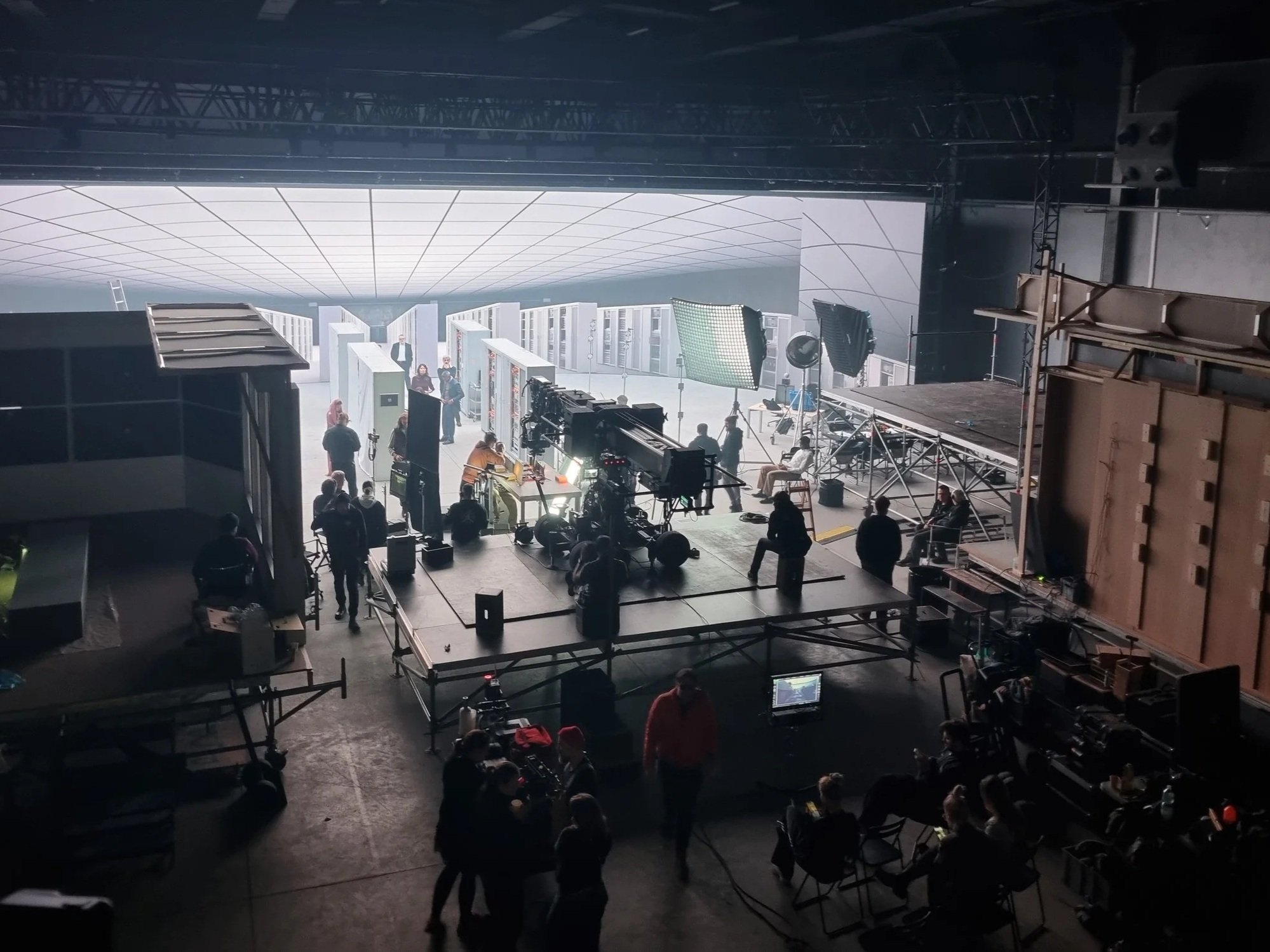 Behind the scenes of a film or photo shoot, showing crew members, lighting equipment, and camera setup in a large studio with a white backdrop and control monitors.