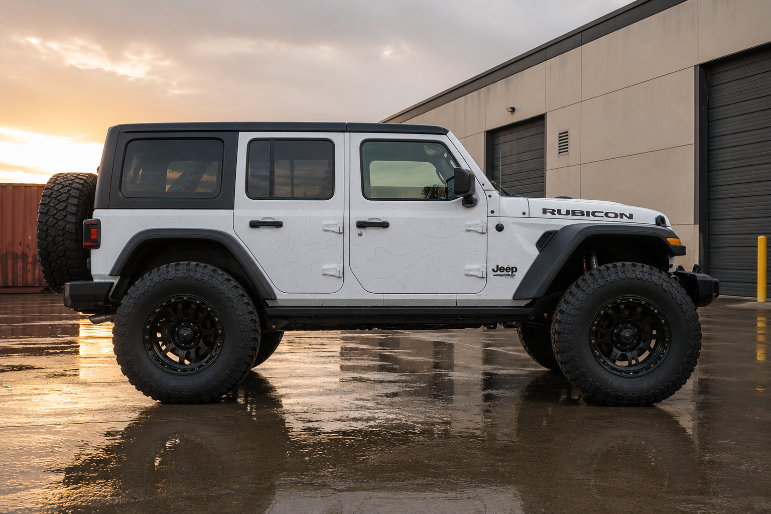 A white Jeep Rubicon Wrangler parked on a wet concrete surface at sunset, with a black spare tire mounted on the rear and a large black off-road tire on the front