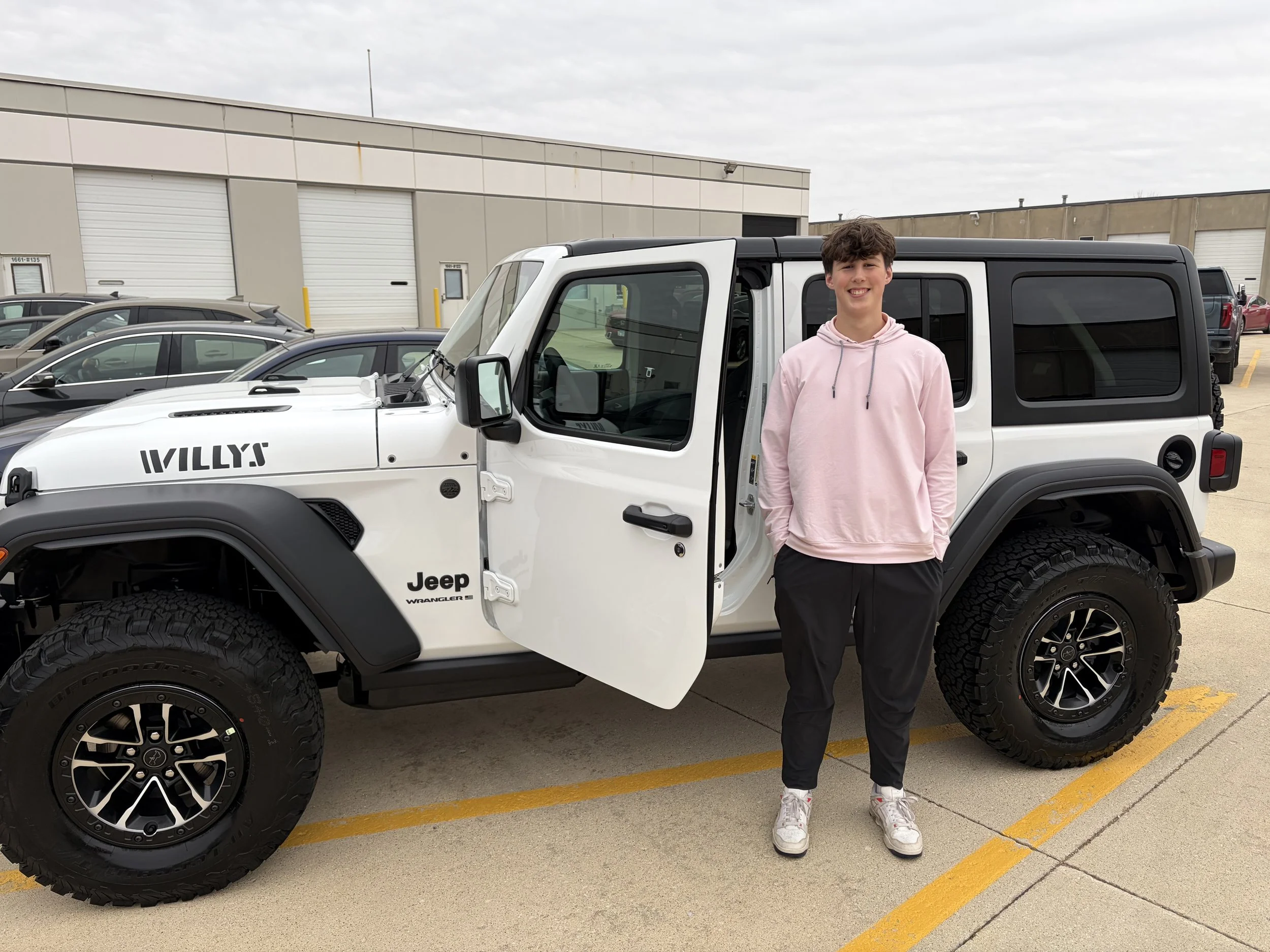 Young man standing next to a white Jeep Wrangler in a parking lot with a large industrial building in the background. The man is smiling and wearing a pink hoodie, black pants, and white shoes.