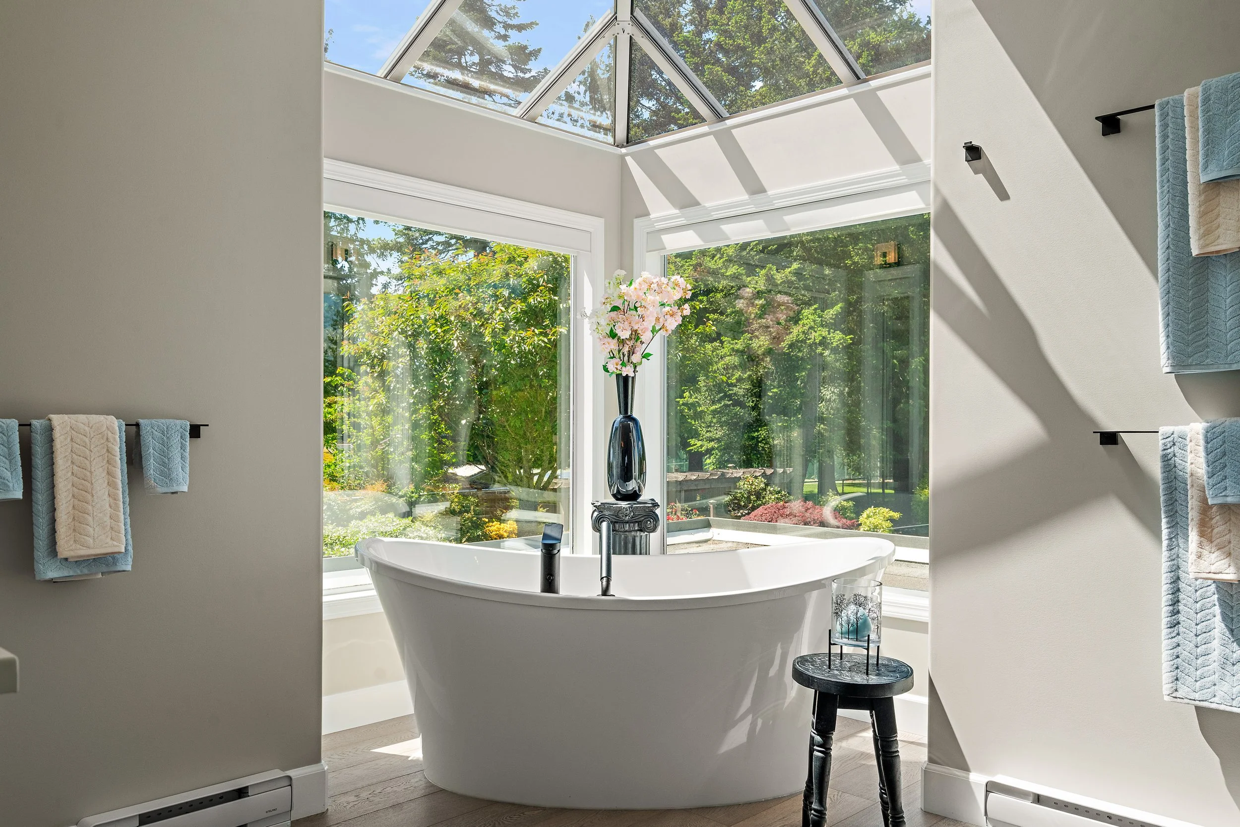 A bright bathroom with a white freestanding bathtub positioned in front of large windows and a glass ceiling, showing a lush outdoor garden. A tall black vase with pink flowers sits behind the tub. Towel racks with blue and beige towels are mounted on the wall, and a small round table with decorative items is near the tub.