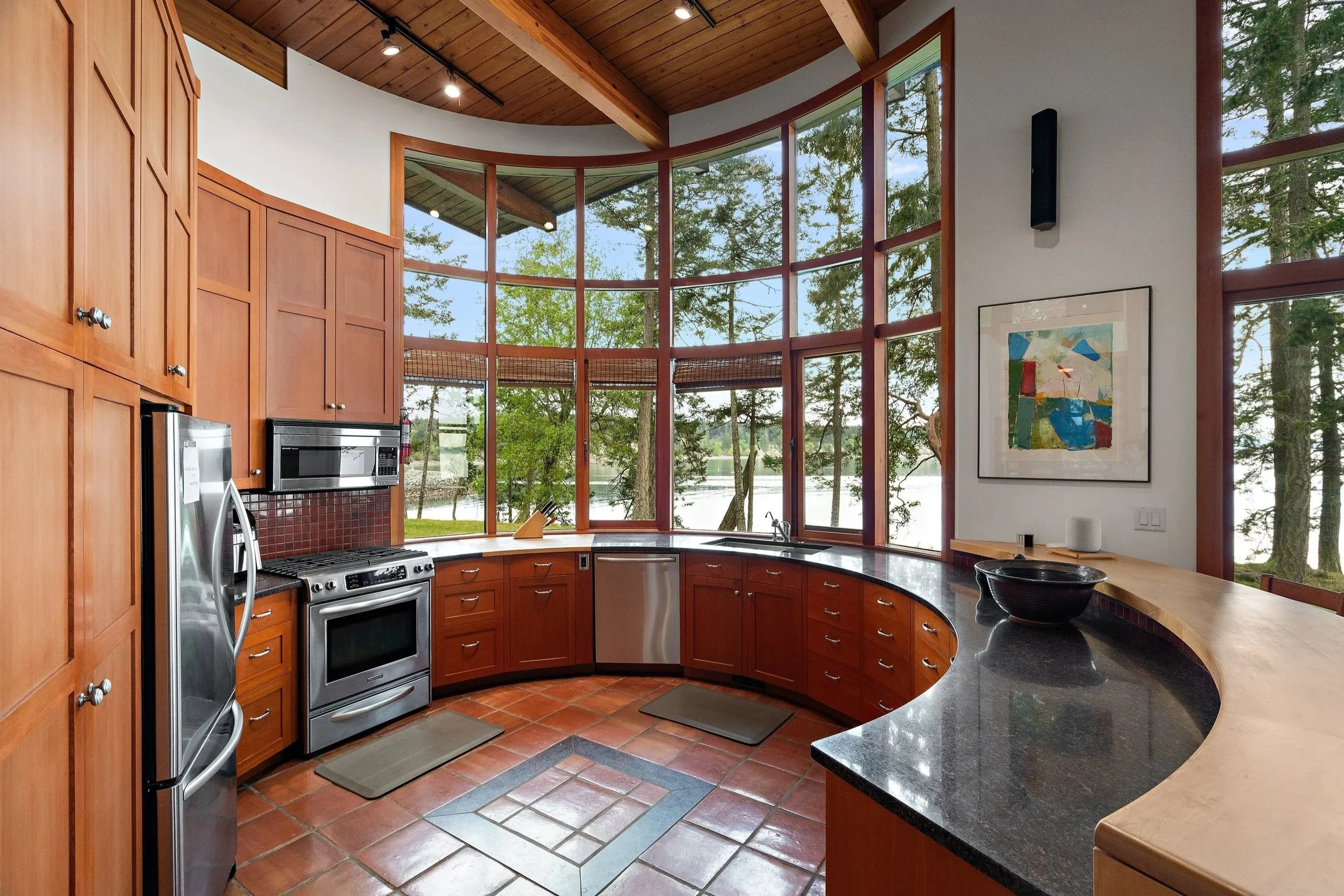 A spacious kitchen with wooden cabinets, stainless steel appliances, black countertops, a curved window view of trees and a lake, and a colorful abstract painting on the wall.