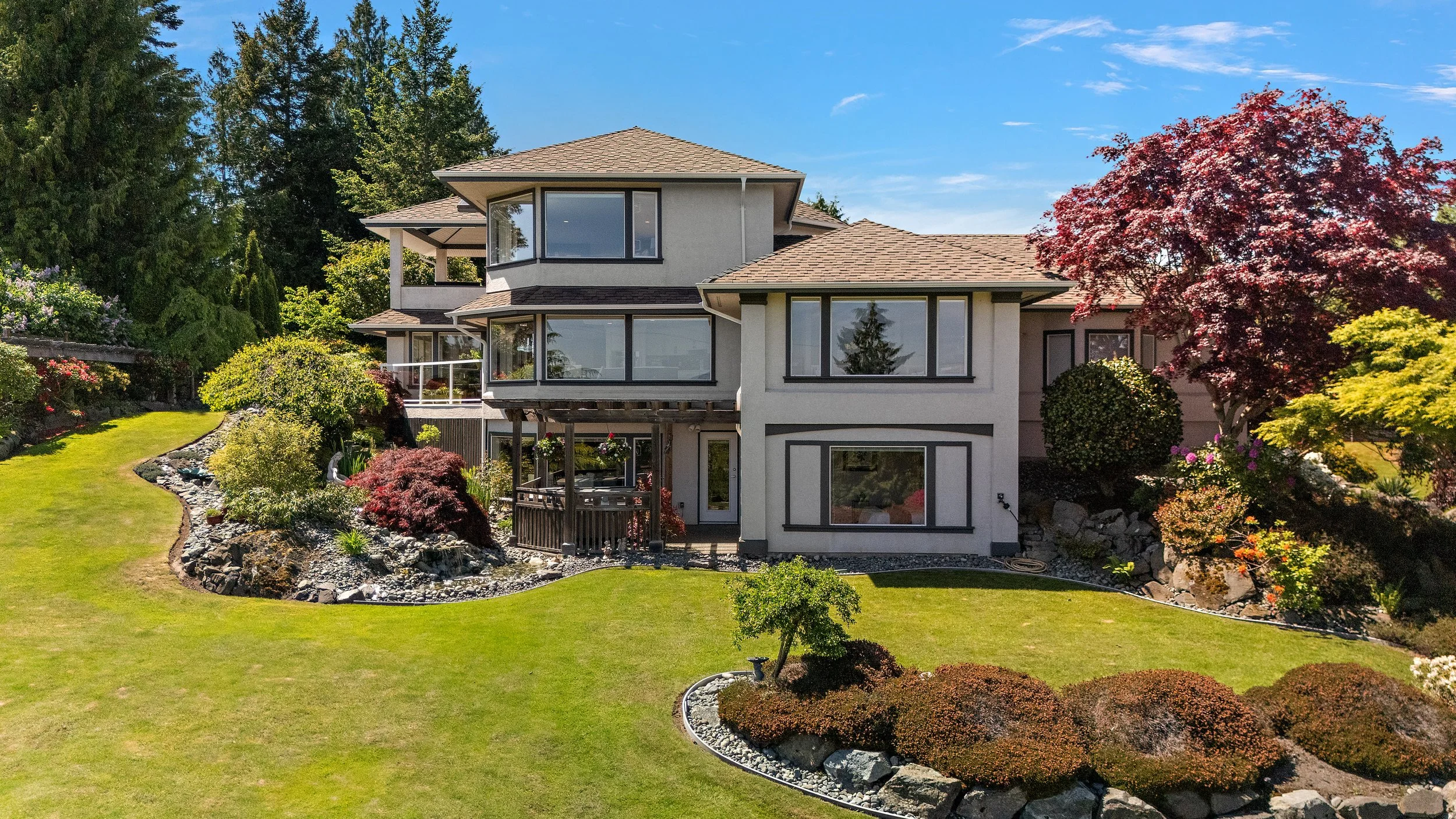 A modern multi-story house with large windows, surrounded by well-maintained garden with various shrubs, colorful trees, and a lush green lawn under a clear blue sky.