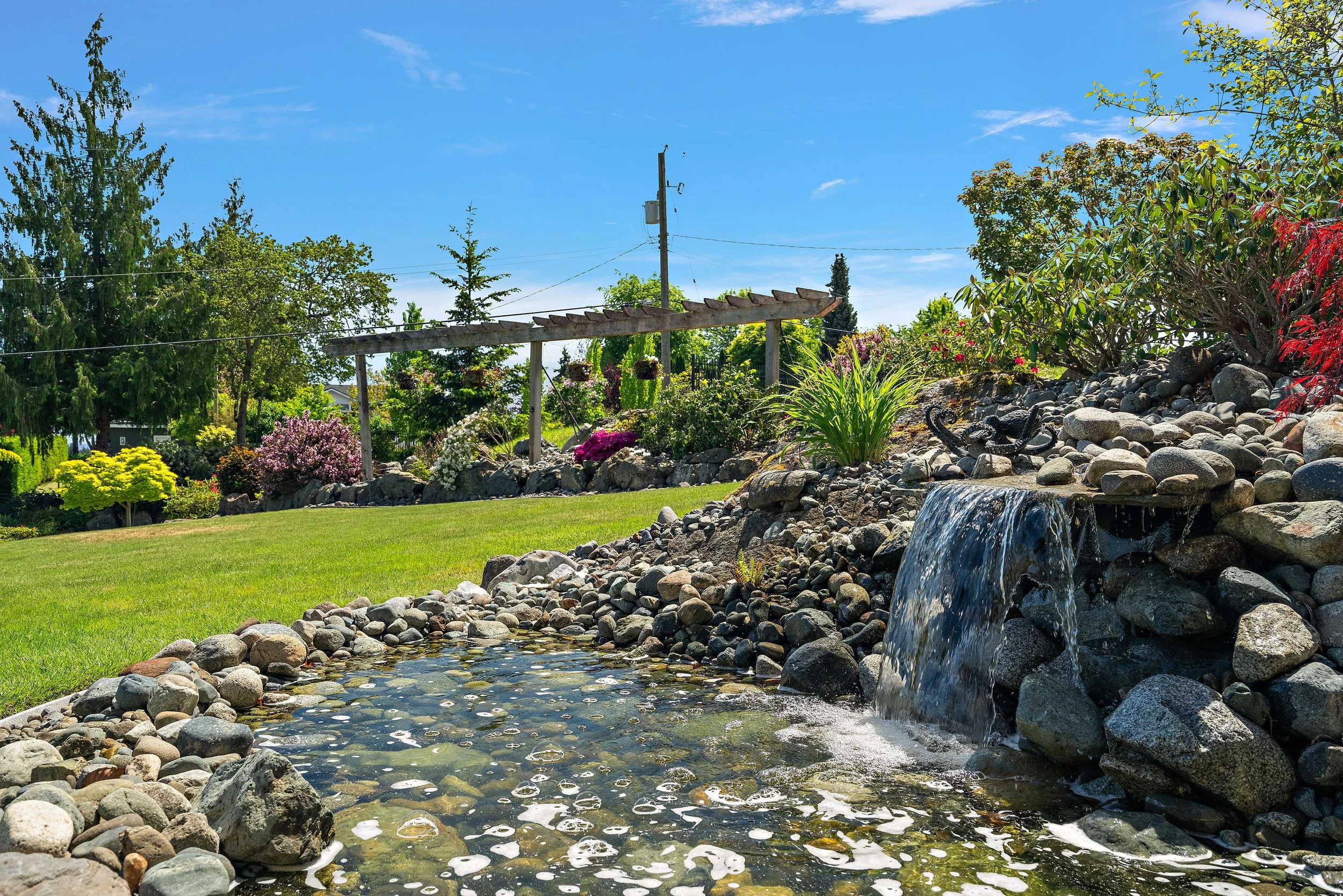 A garden with a small waterfall flowing over rocks, surrounded by lush green trees and bushes, under a clear blue sky.