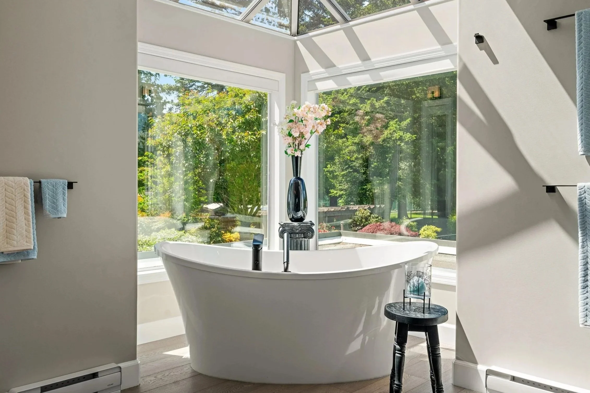 A luxurious bathroom with a standalone bathtub positioned in front of large glass windows and a skylight, allowing natural light to fill the space. The bathroom features a vase of pink flowers on a black pedestal, and there are towels hanging on black towel bars on the wall.