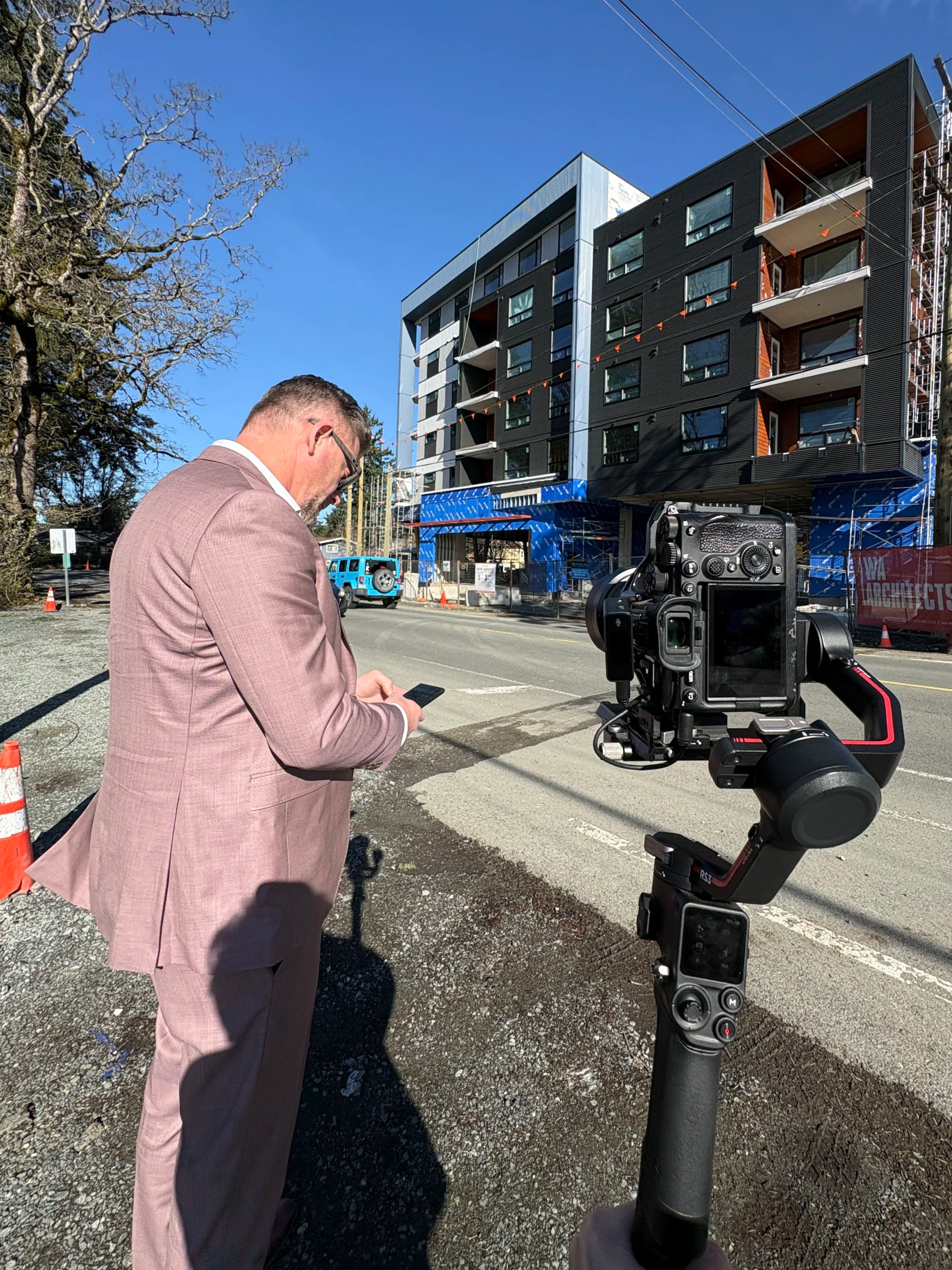 Man in a pink suit looking at his phone in front of a building under construction, with a camera on a stabilizer in the foreground and a clear blue sky.