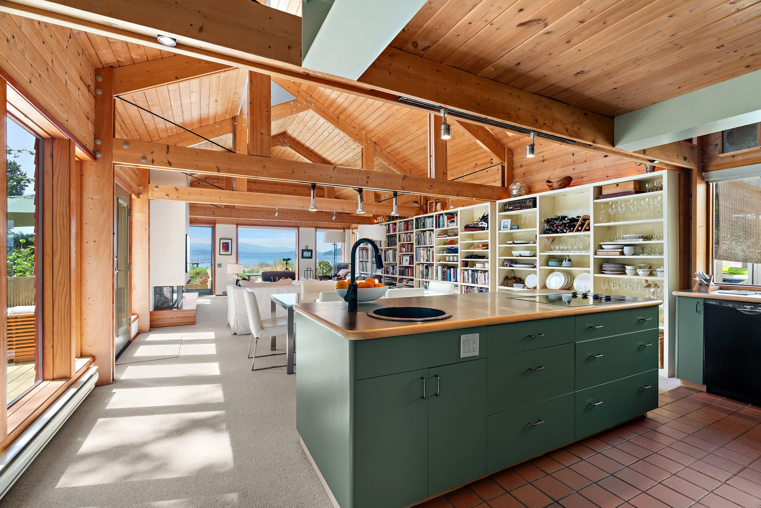 Open-concept kitchen and living space with a vaulted wood ceiling, large windows showing outdoor scenery, a green kitchen island with a built-in sink, open shelving with dishes and glassware, and a dining table with white chairs.