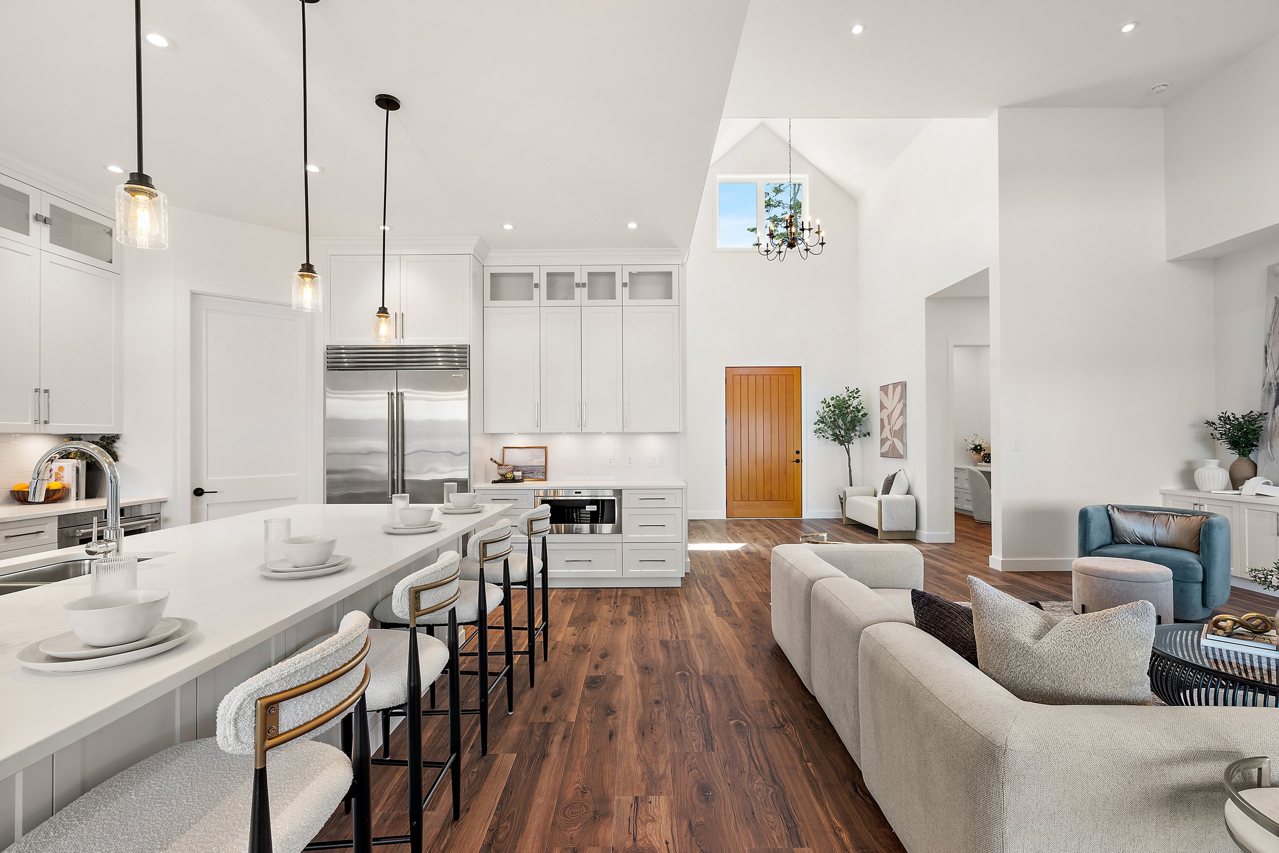 Open concept living room and kitchen with white walls, wooden flooring, modern and traditional light fixtures, including a chandelier and pendant lights, kitchen with white cabinetry, stainless steel refrigerator, island with white counter and place settings, and seating area with beige and blue furniture.