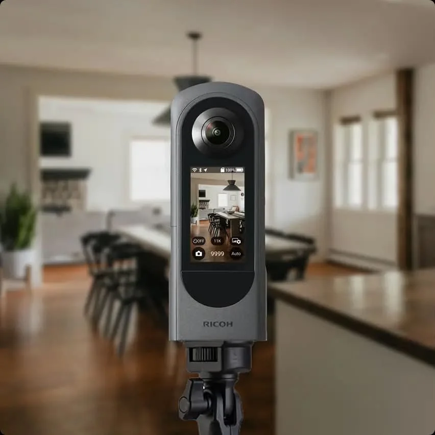A camera mounted on a tripod capturing an indoor dining area with a long table, chairs, windows, and a hanging light fixture.