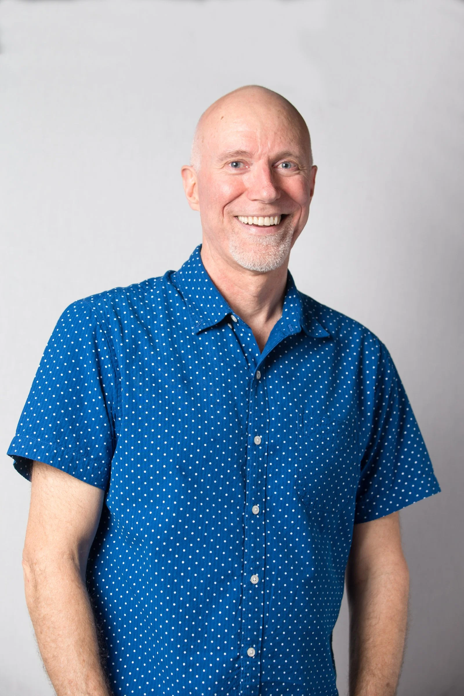 A middle-aged man with a bald head, blue eyes, and a white goatee, wearing a blue shirt with white polka dots, smiling, against a plain, light gray background.