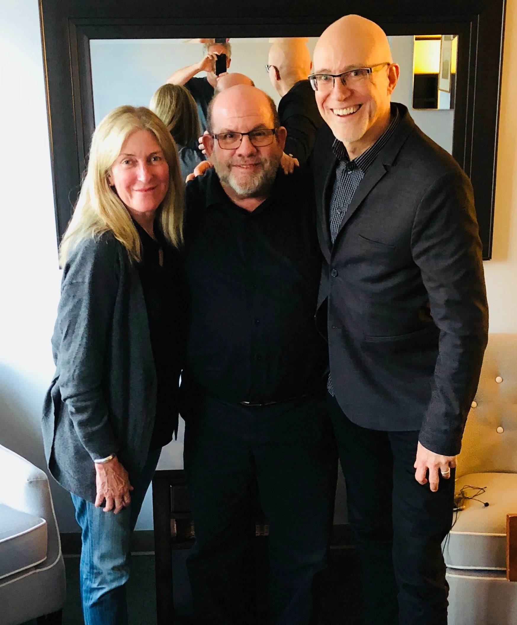 Three people are smiling and posing for a photo in a room with a mirror behind them. The mirror reflects a man taking the photo with a phone. The three people are a woman with long blonde hair and a man with glasses and a beard, and another man with a bald head and glasses wearing a suit. There is a lamp and some furniture visible in the background.