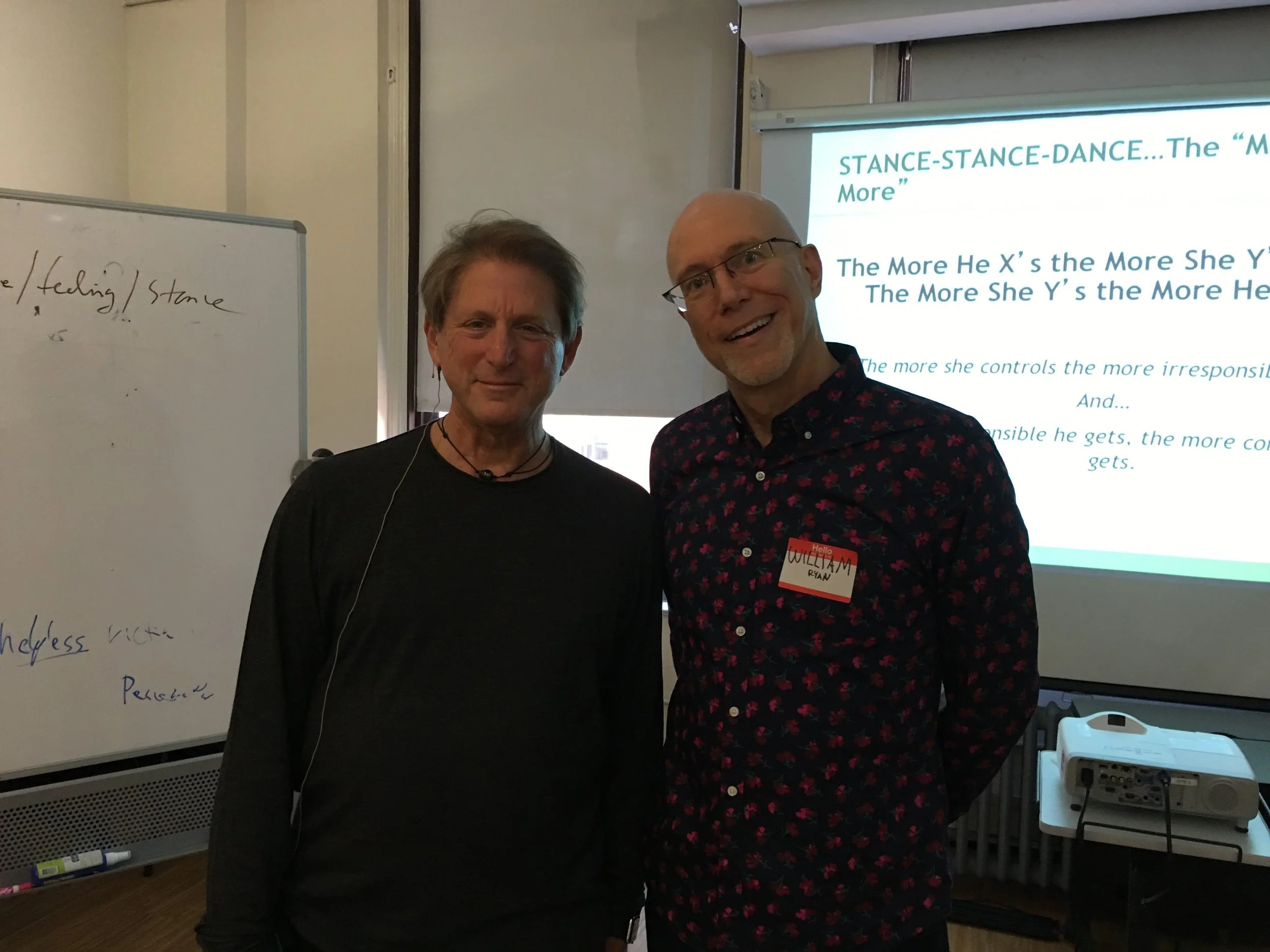 Two men smiling in a classroom with a whiteboard and a projector screen in the background. One man is wearing a dark shirt, the other a floral shirt with a name tag that says William Rian.