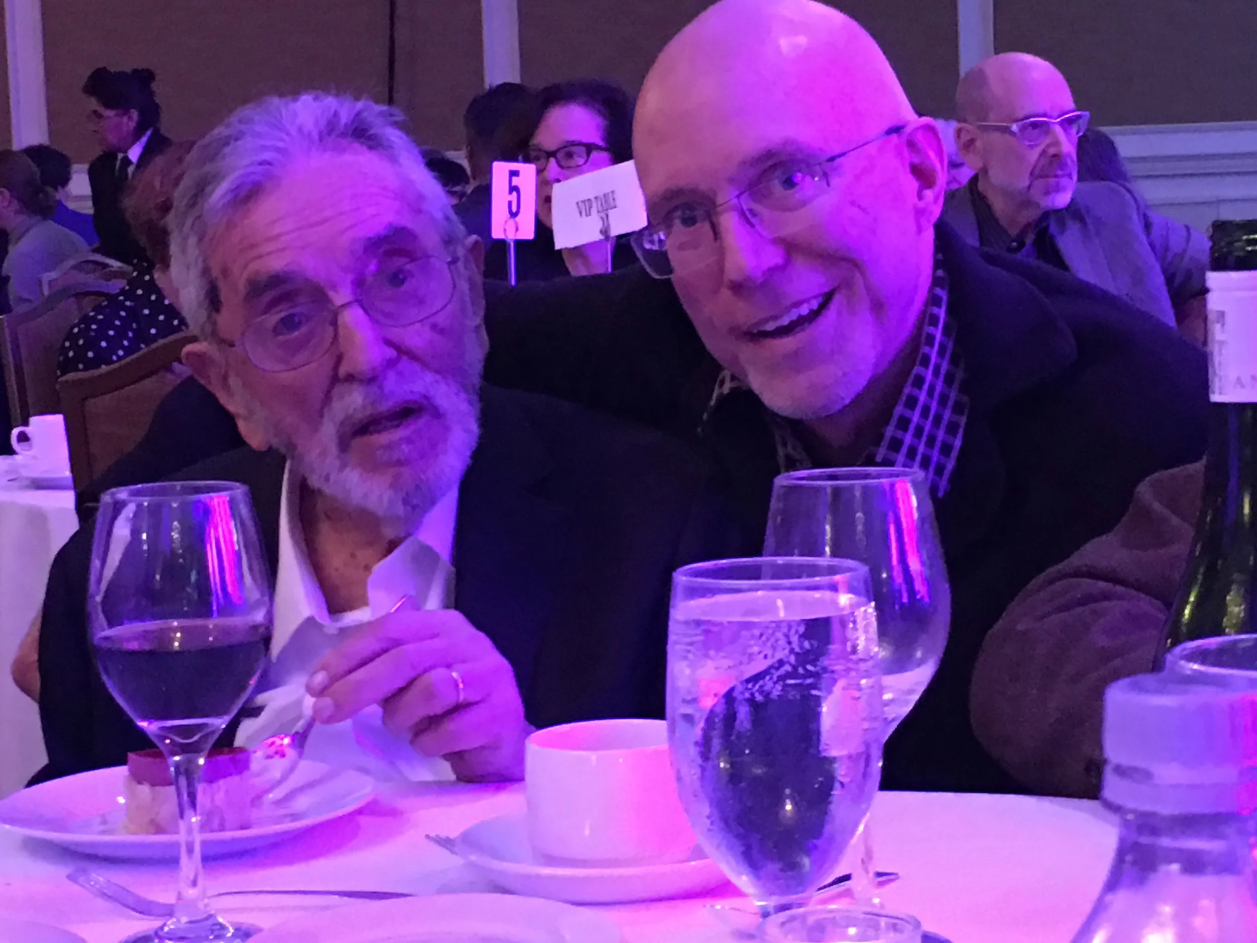 Two older men sitting at a table, holding glasses of wine. They are at a dinner event with purple lighting, and there are other people in the background, including a woman wearing glasses. The table has wine glasses, a coffee cup, and plates.