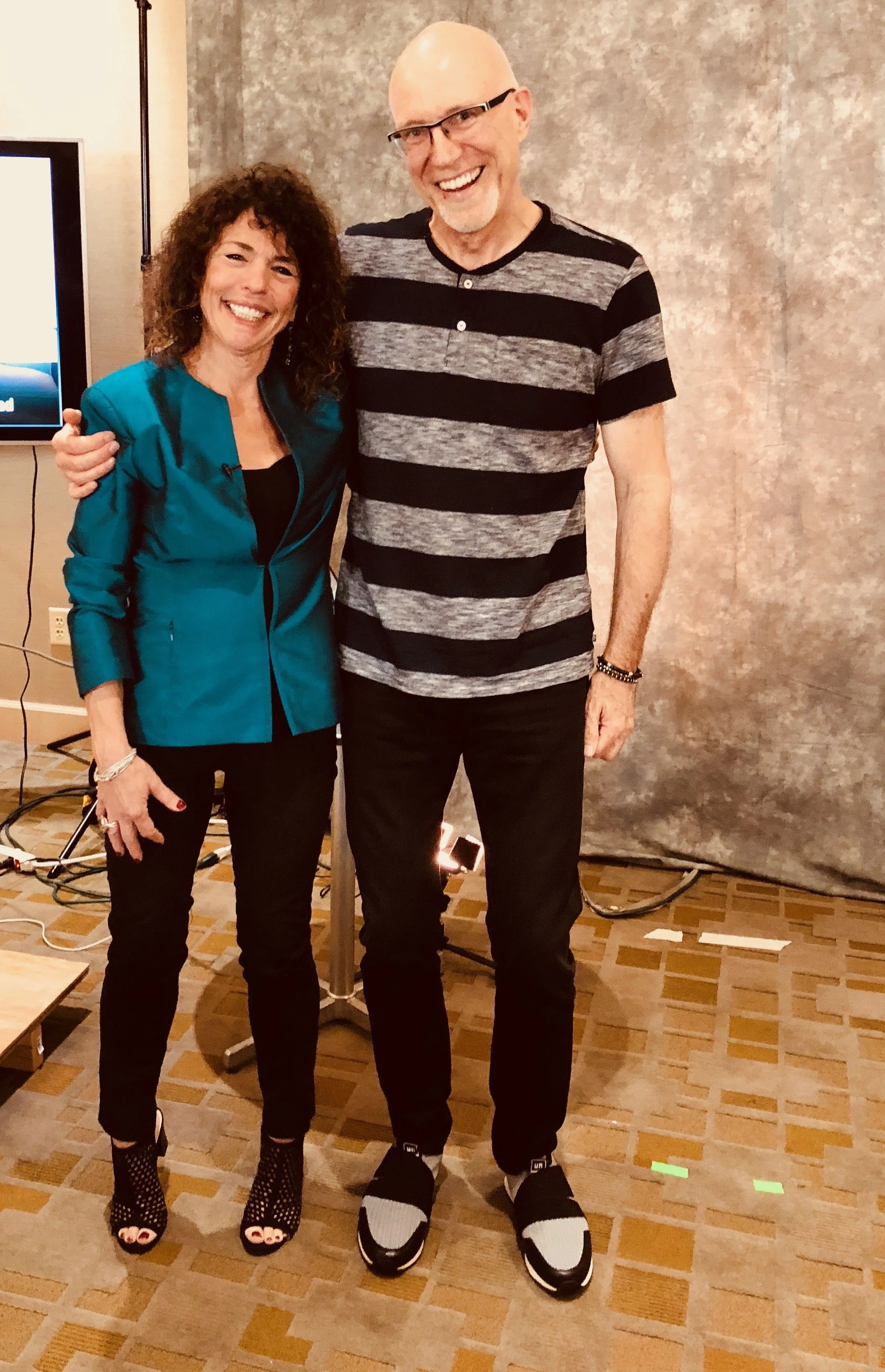A man and a woman standing close together, smiling for the camera in an indoor setting with a textured wall behind them. The woman is wearing a teal jacket, black pants, and high-heeled shoes with cuts and patterns, and the man is dressed in a striped black and gray T-shirt, black pants, and sneakers.