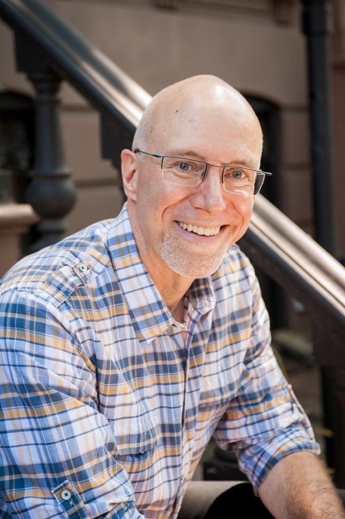 A smiling man with glasses and a short beard, wearing a plaid button-down shirt, sitting outdoors near a staircase.