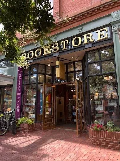 The storefront of a bookstore called 'BOOKSTORE' with large glass windows and open doors, located on a brick-paved sidewalk with a bicycle parked outside and greenery around.