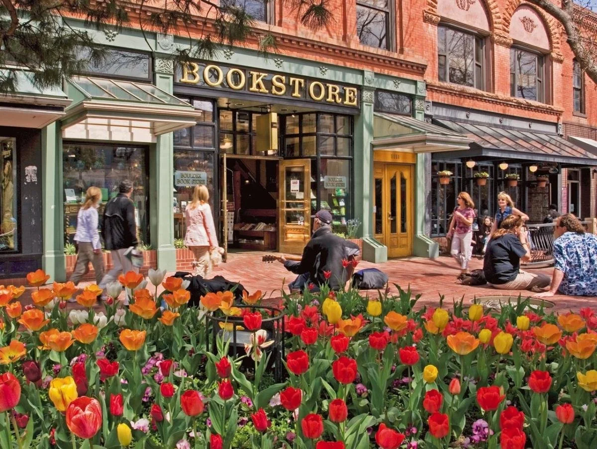 A bookstore with a green storefront and a yellow sign that reads 'BOOKSTORE.' There are people walking and sitting outside, some with musical instruments. The foreground has a bed of colorful tulips.