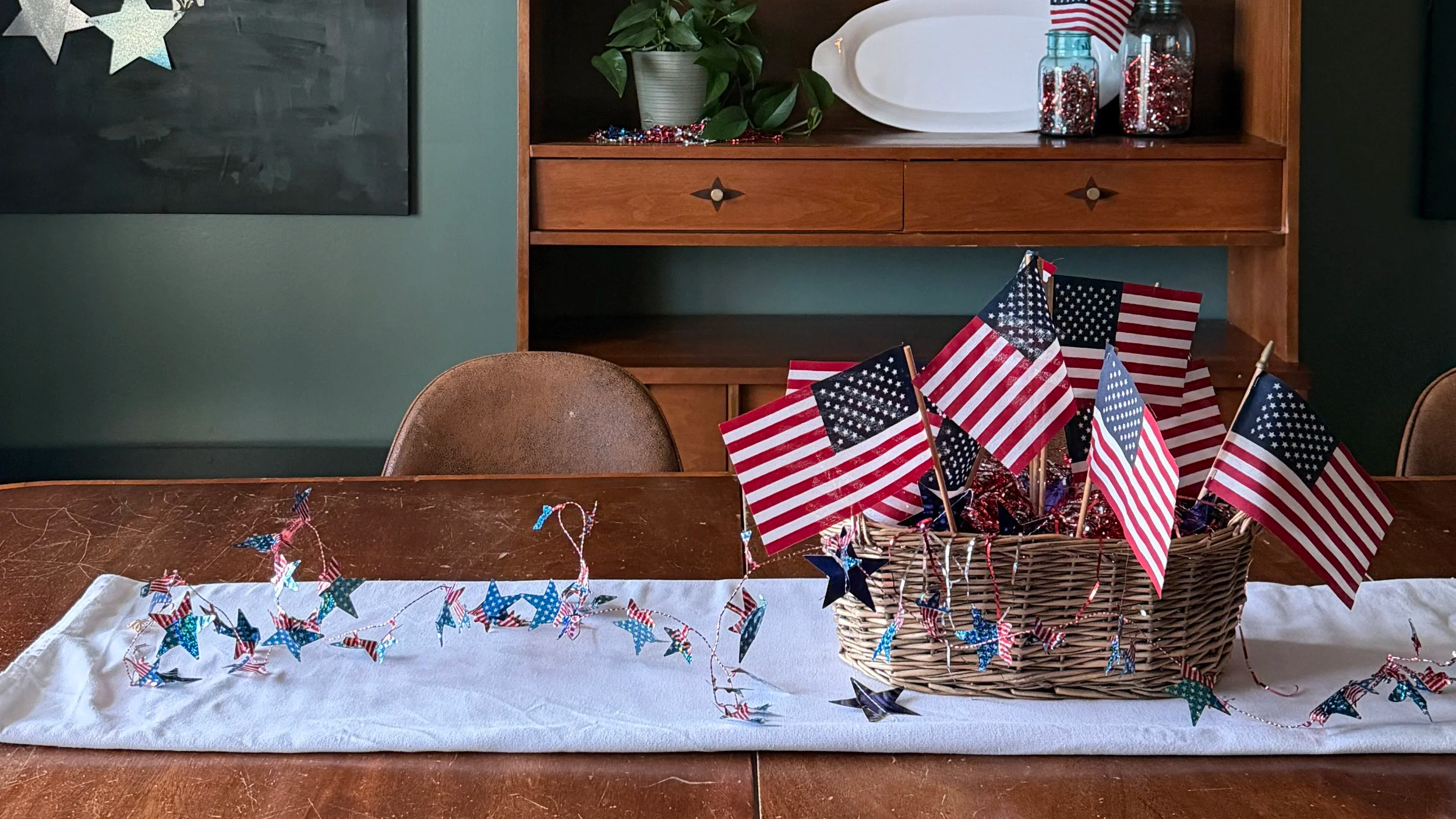 A warm, deep green dining room with mid century furniture decorated for America's 250th birthday. A white table runner, brown woven basket with American flags and tinsel in it.