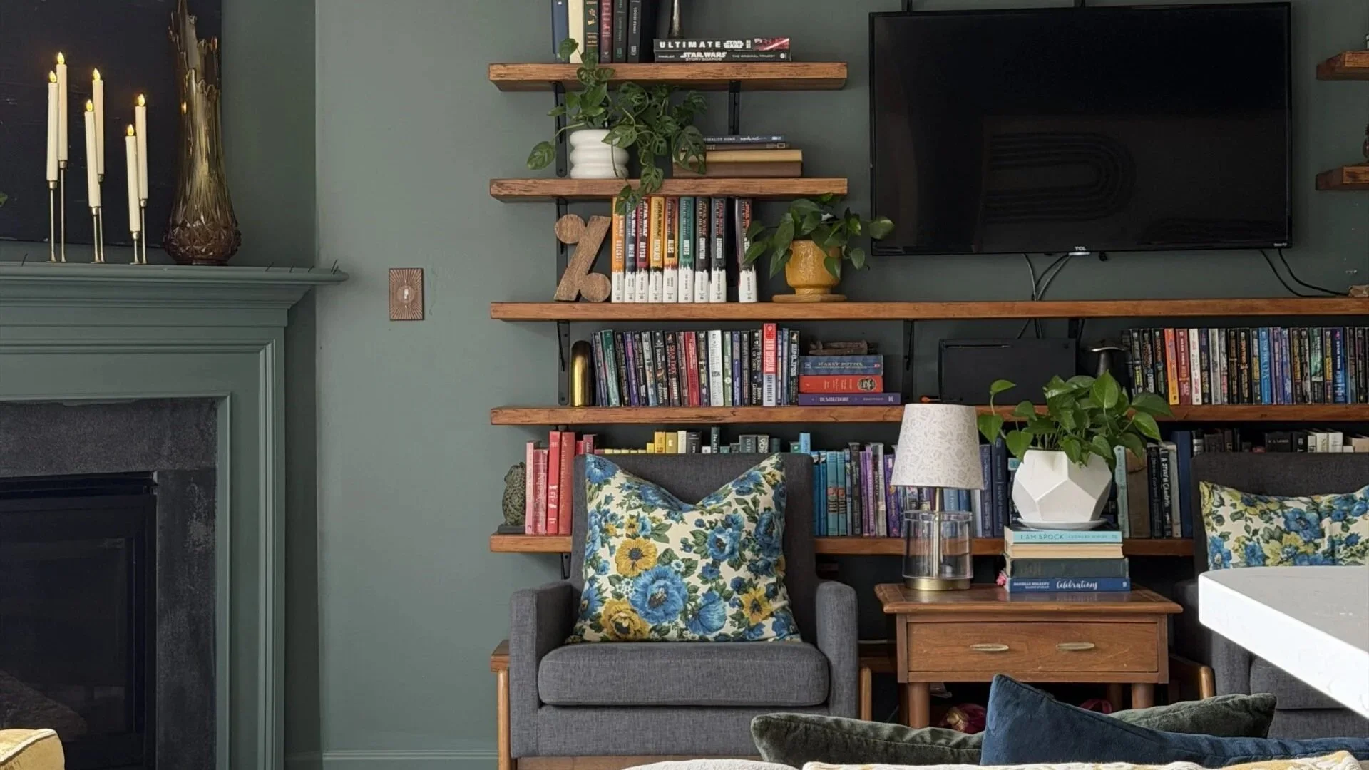 Dark green living room with warm wood shelves surrounding a tv mounted on the wall. The vibe is academia with a side of mid century modern, but make it cozy. there are chairs  and a table in front of the wall of shelves, the table has a lamp on it.