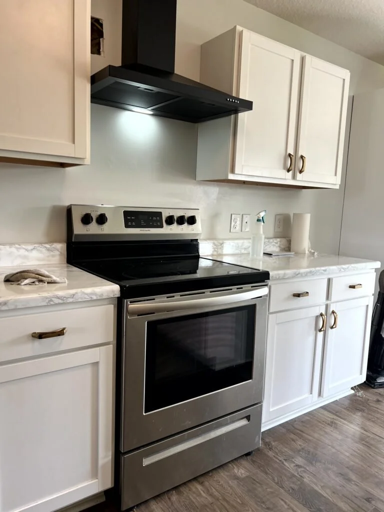  Kitchen with white cabinets the microwave and over the microwave cabinet gone, a new black range hood, and a hole in the wall left by the original contractors who intially put the outllet in the wrong place.  