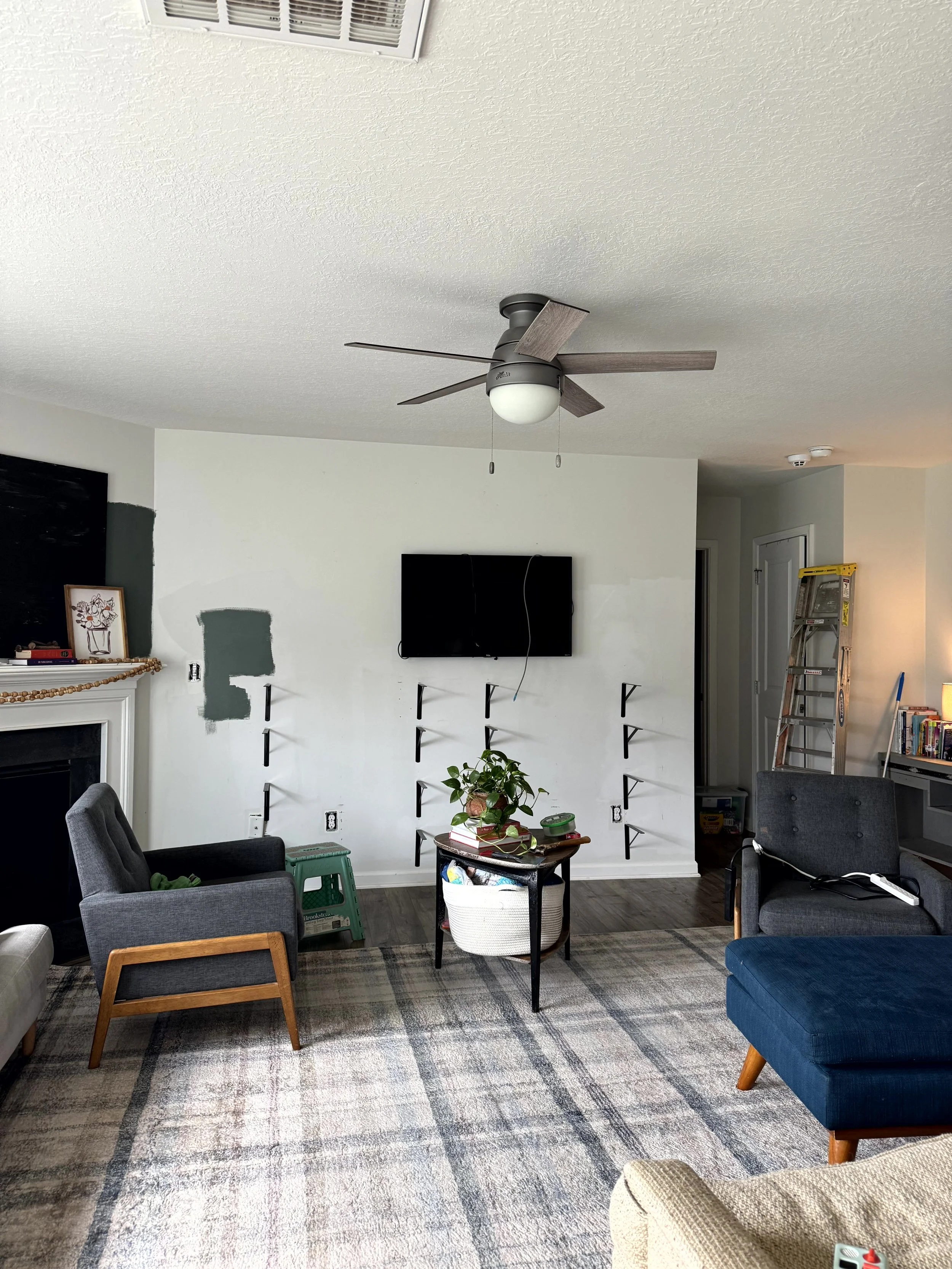  A progress picture of a living room that is currently gray but has a large green swatch of paint on multiple walls. The shelves have been removed but the brackets are still there, there is a ladder leaning against the wall and the chairs and table h