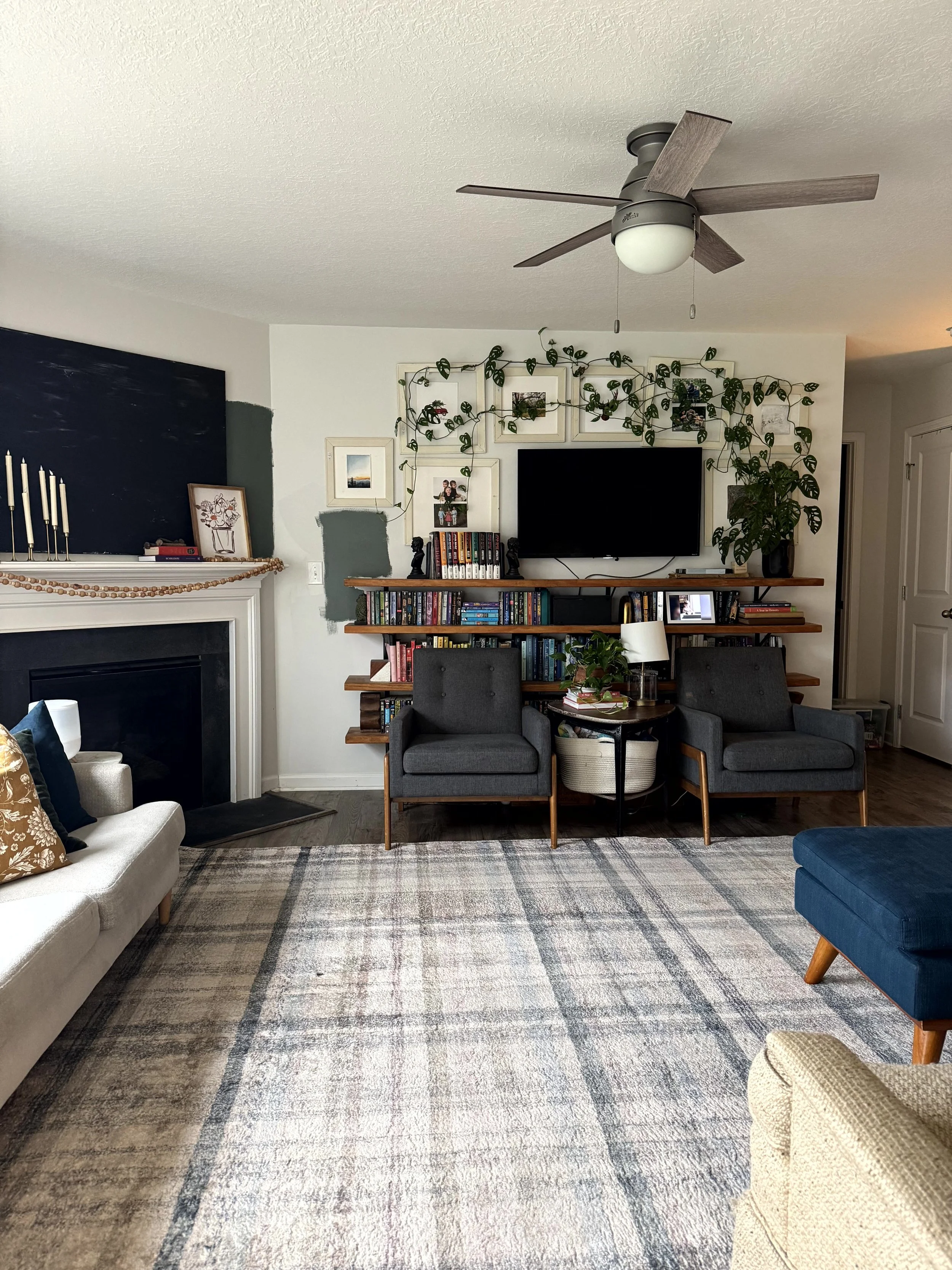  The before image for this living room makeover. Light gray walls, with warm wood shelves partially up the wall surrounding the TV. Chairs in front of the shelving, with a table in between. A warm patterned rug with shelves of books and a plant climb