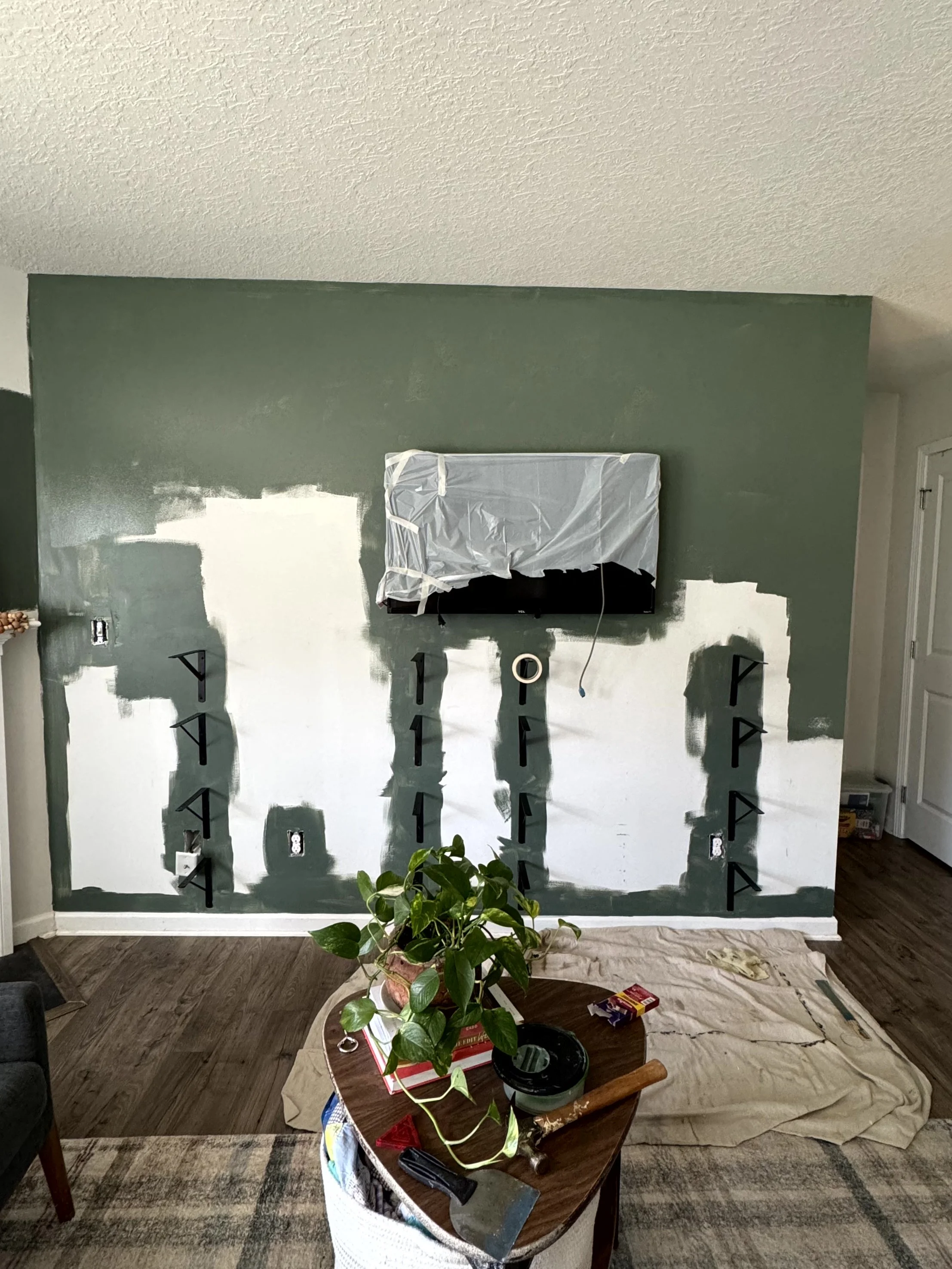 A partially painted wall, part green part gray, the TV is covered in a garbage bag to protect it and there is a drop cloth laid out on the floor to catch drips.  