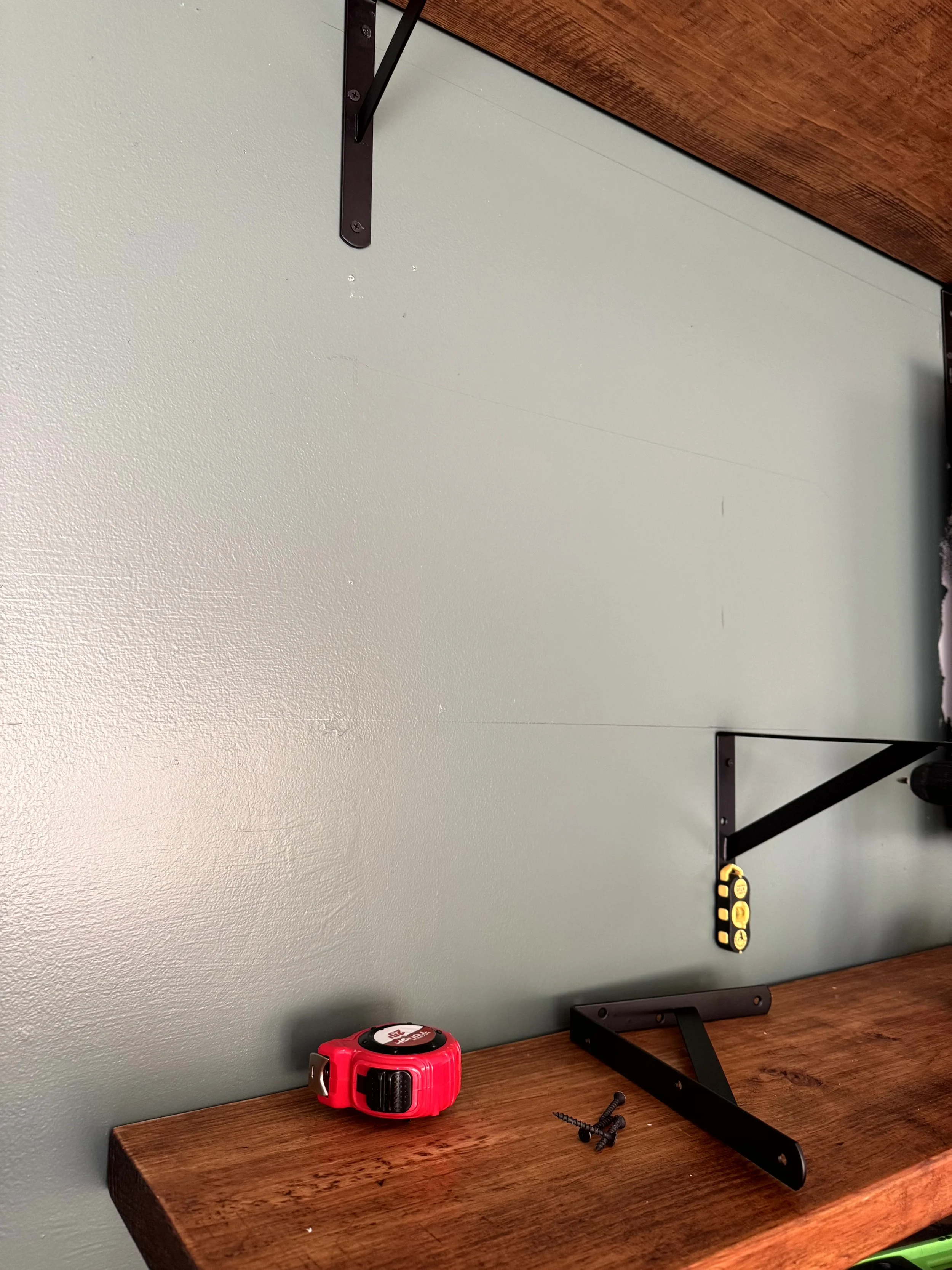  A lose up of the green walls, and brackets and shelves. A magnetic stud finder is attached to one of the brackets that is being installed  to make sure it is level. There are other brackets, screws, and a tape measure laying on the shelf below.  